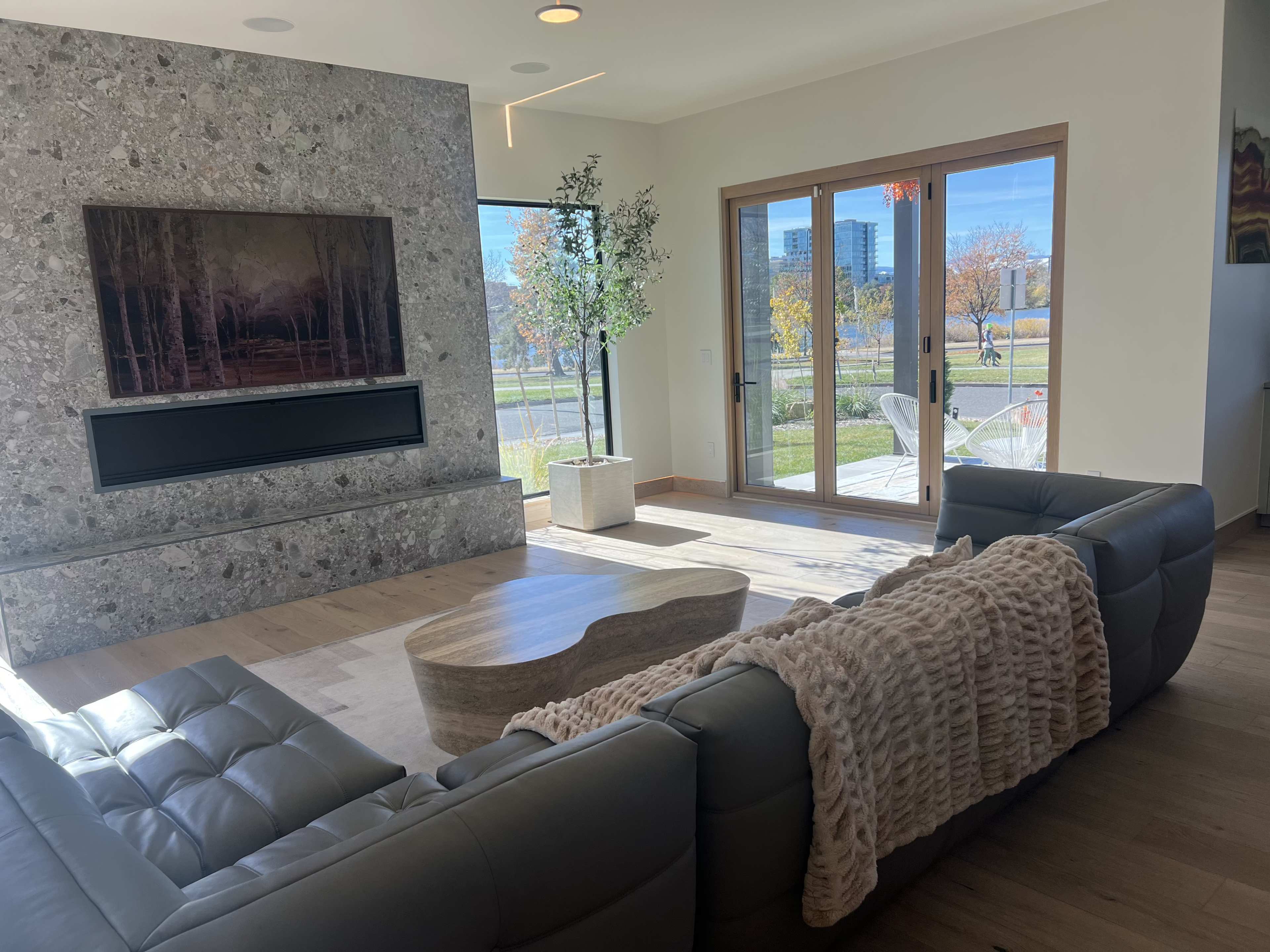 The image shows a modern living room with a stone fireplace, large windows, and a sleek gray sofa adorned with a cozy throw blanket.