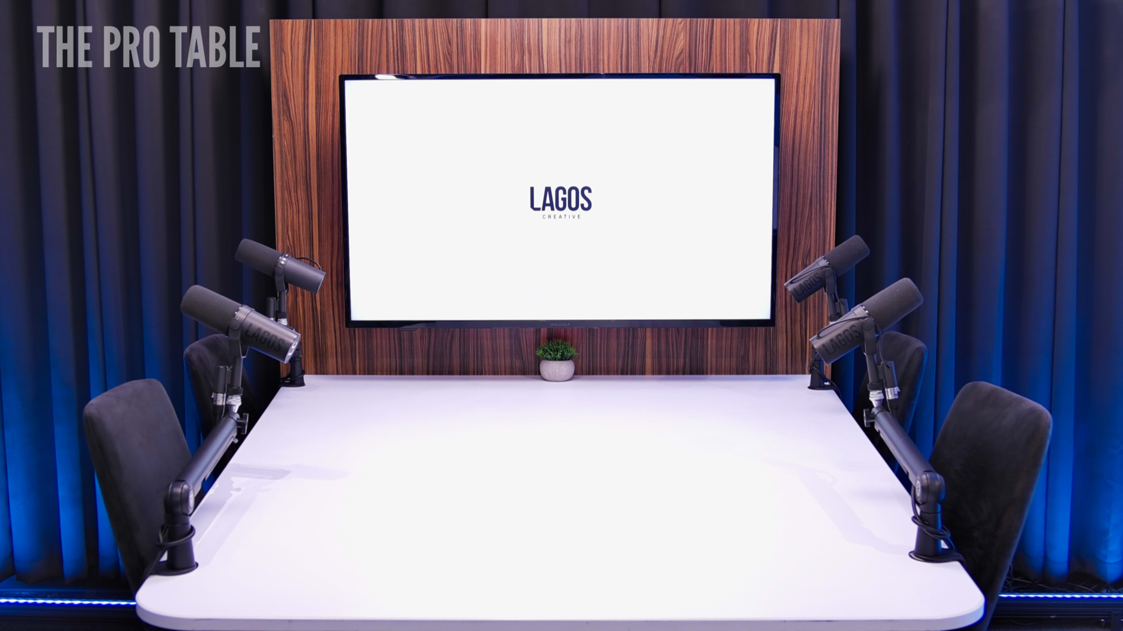 A modern podcast studio features a large screen displaying "LAGOS" on a wooden backdrop, with microphones positioned at two sides of a white table.