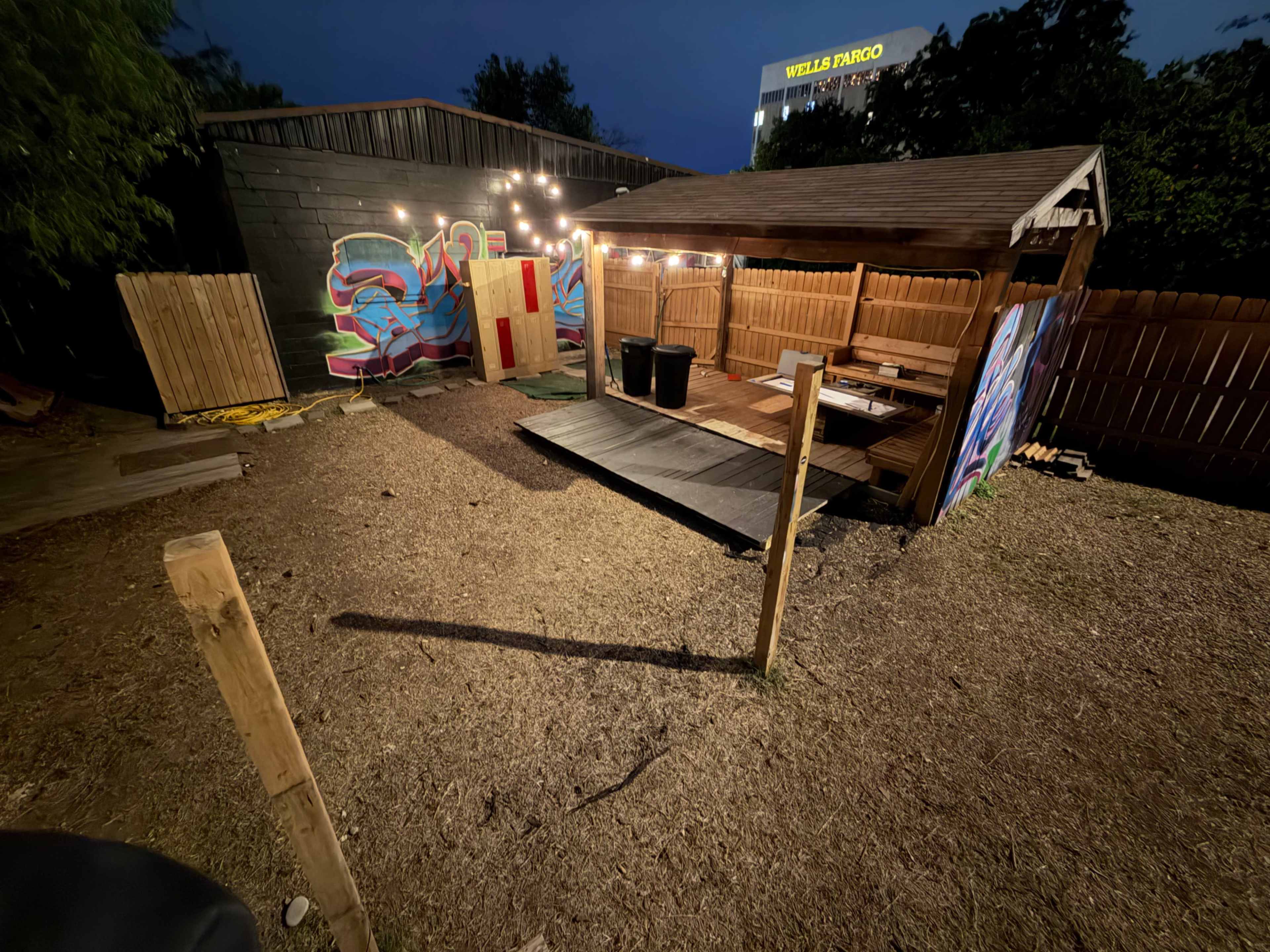 The image shows a dimly lit backyard with a wooden structure resembling a gazebo, surrounded by a wooden fence and featuring a vibrant mural on one wall.