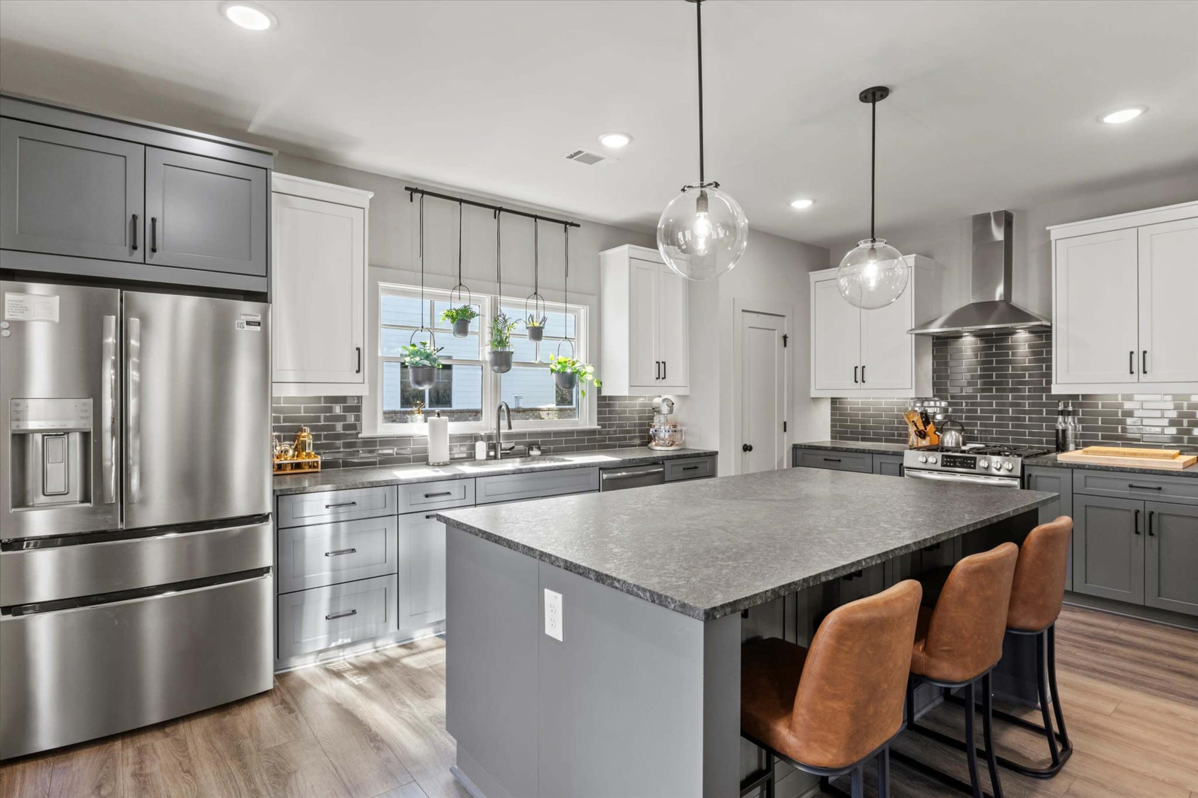 The kitchen features a central island with three bar stools, sleek cabinetry in gray and white, a stainless steel refrigerator, and a window adorned with hanging plants.