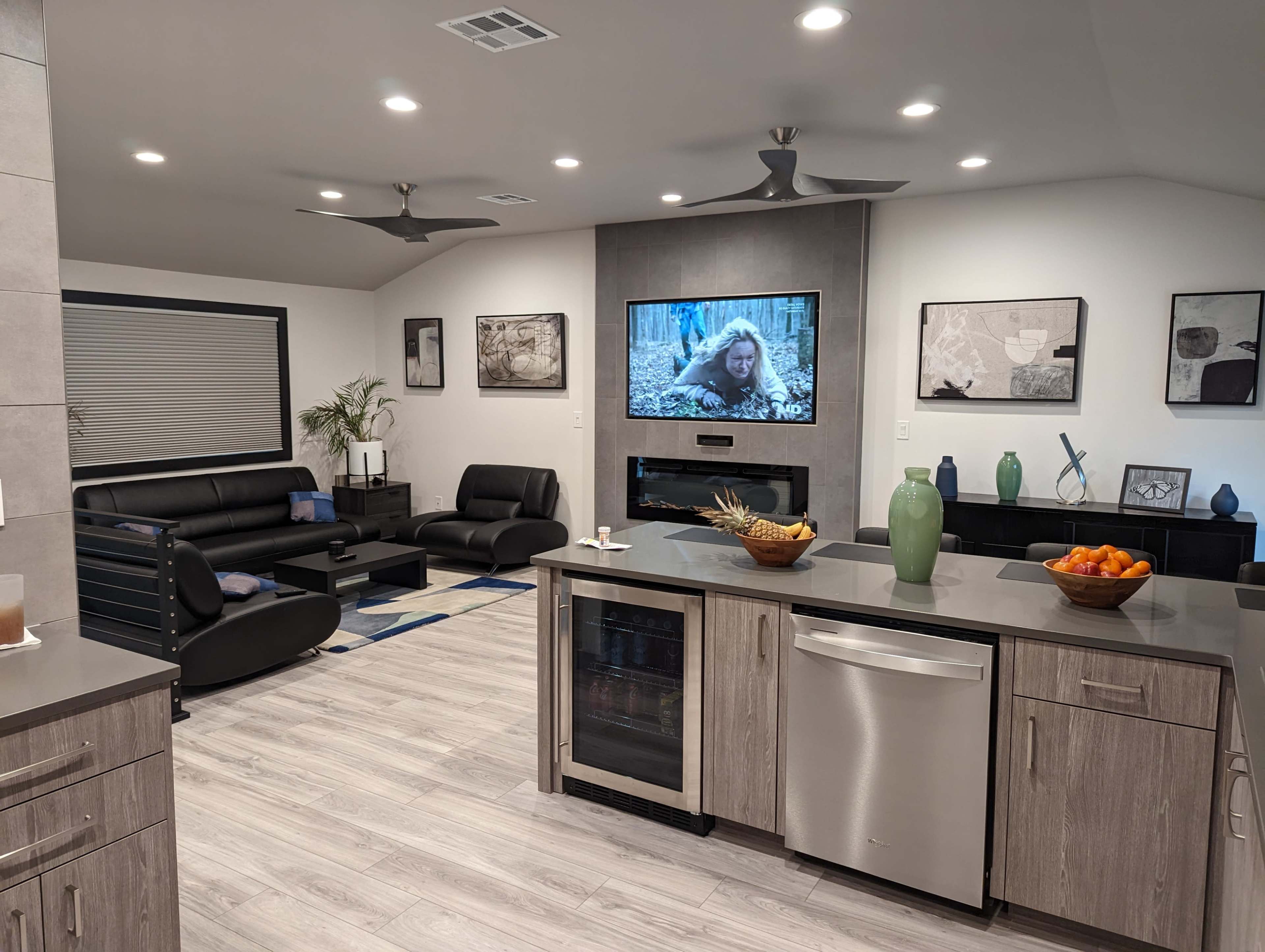 A modern living room and kitchen area featuring dark furniture, a large wall-mounted television displaying a scene from a movie, and a kitchen island with various decorative items and fruit.