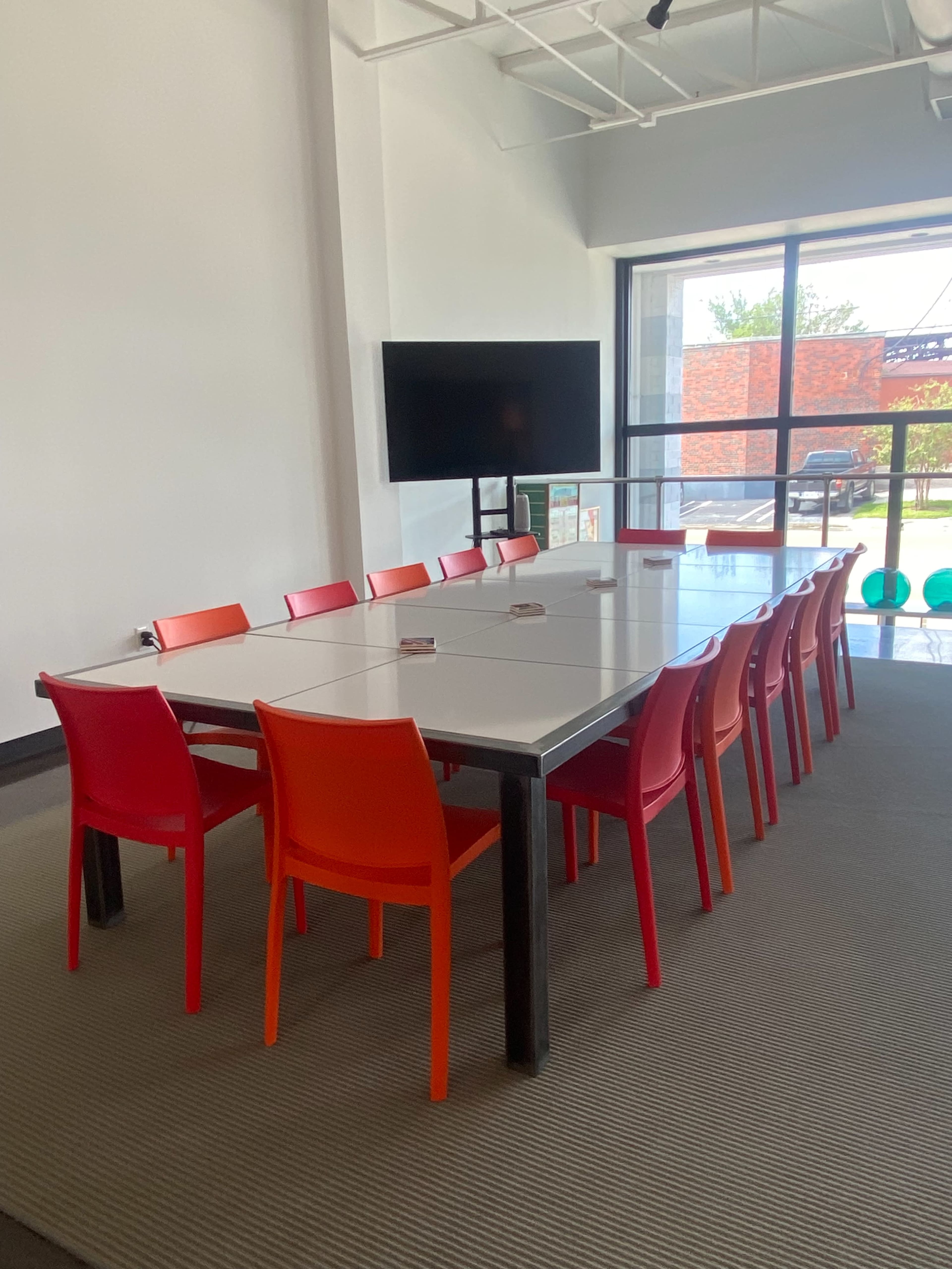 A modern meeting room features a long table surrounded by red and orange chairs, with a large screen mounted on the wall and windows providing natural light.