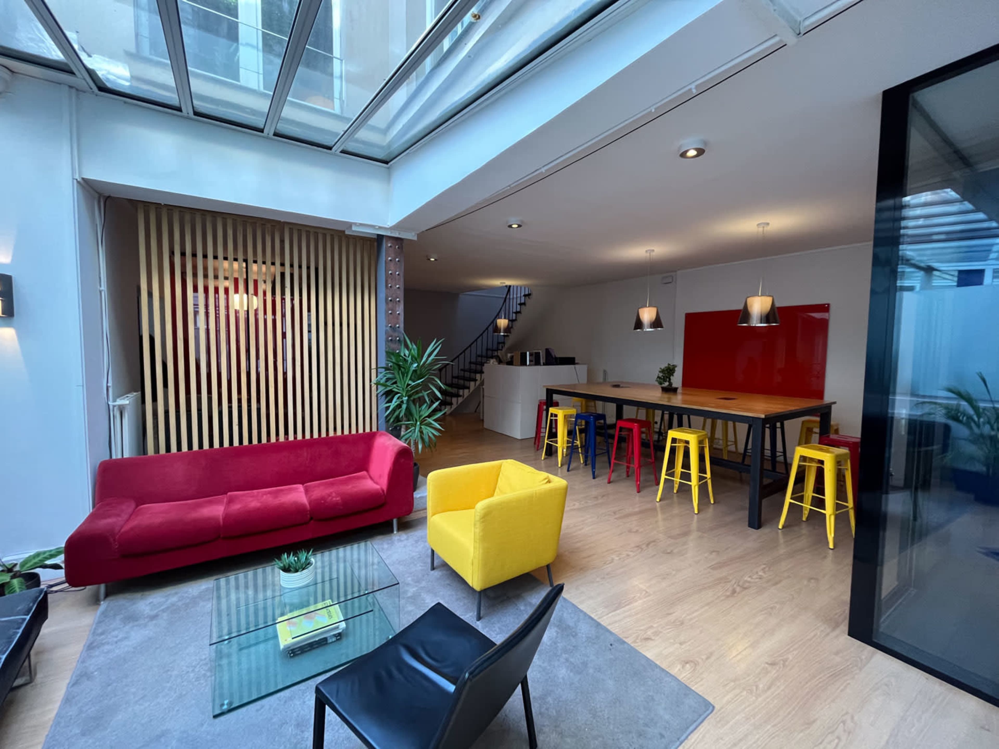A modern, open-concept living space featuring a red sofa, a yellow chair, a glass coffee table, and a dining area with colorful stools.