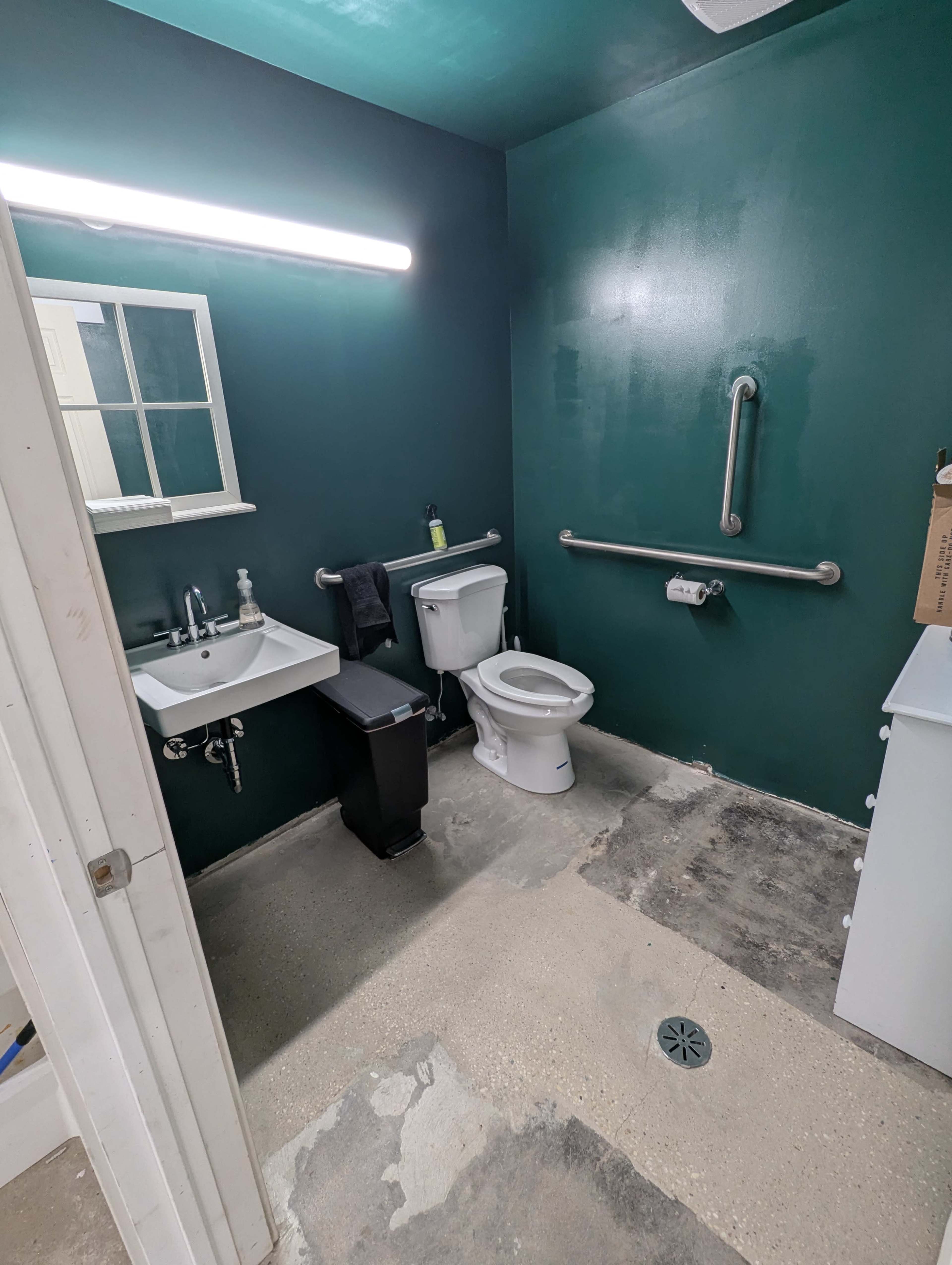 The image shows a bathroom with green walls, a toilet, a sink, a grab bar, and a partially finished floor.