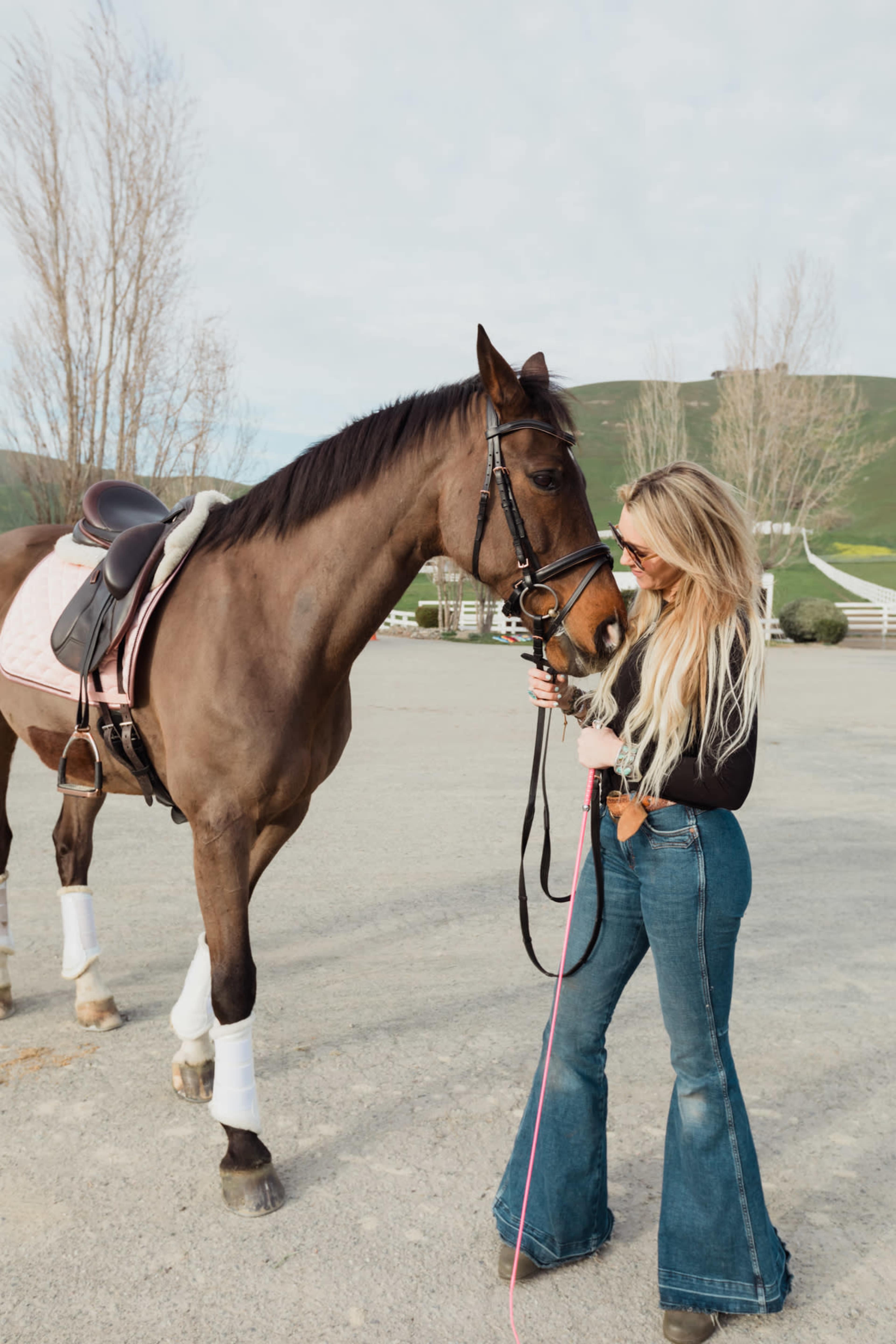 A woman in flared jeans gently interacts with a brown horse in an outdoor arena.