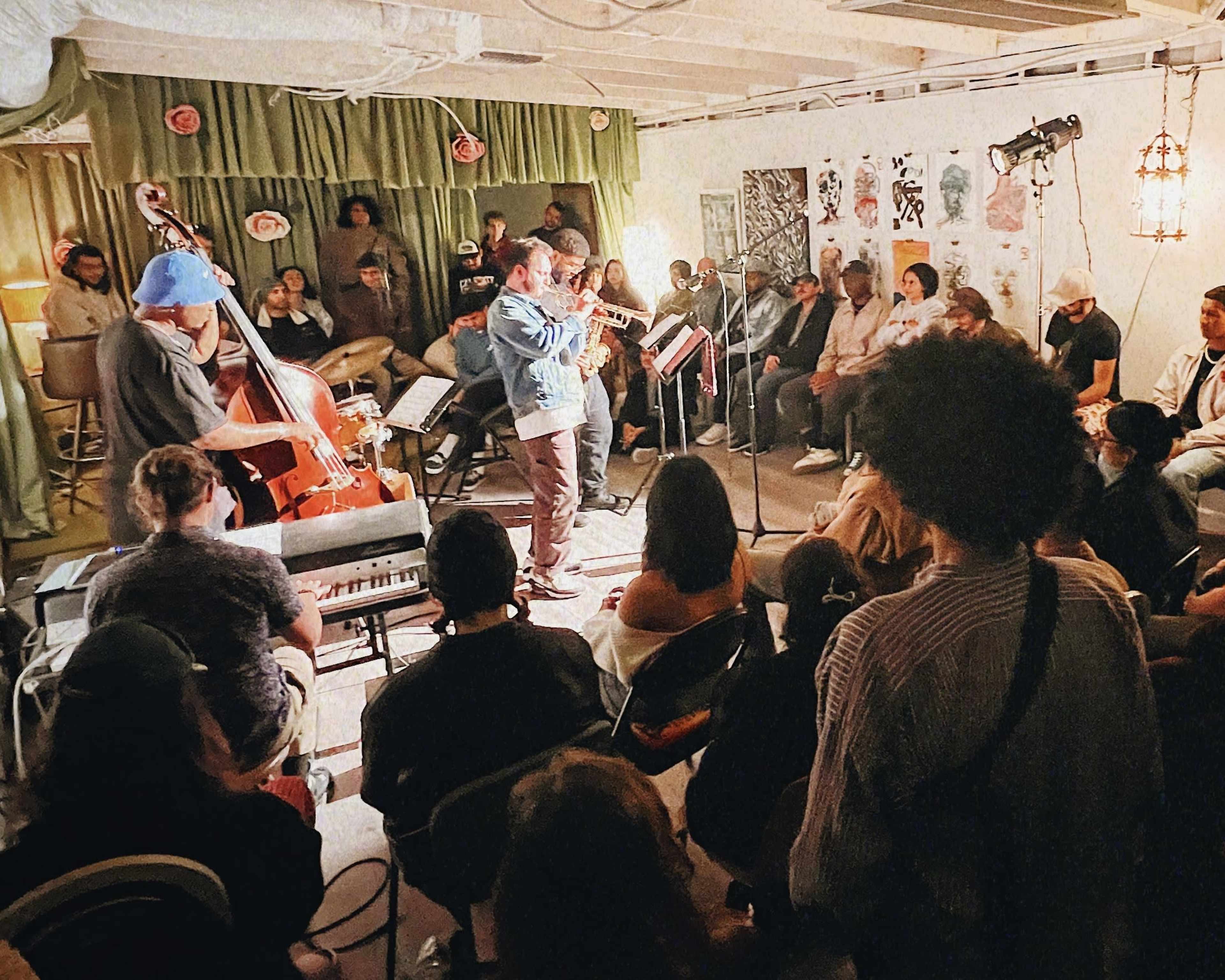 A group of musicians performs on a small stage in a cozy room filled with an audience seated in chairs.