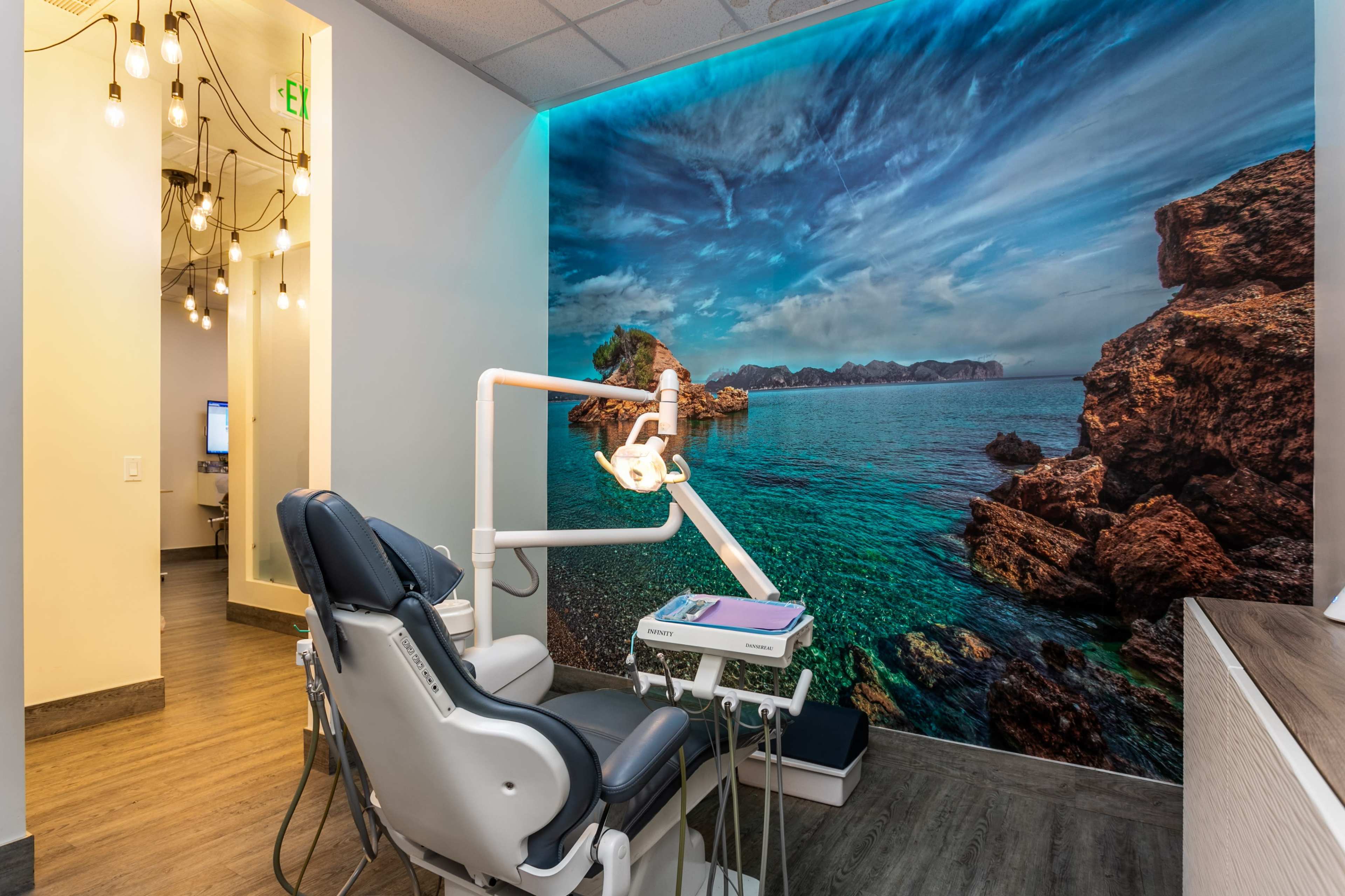 A dental treatment room featuring a dental chair, an overhead light, and a large mural of a coastal landscape on the wall.