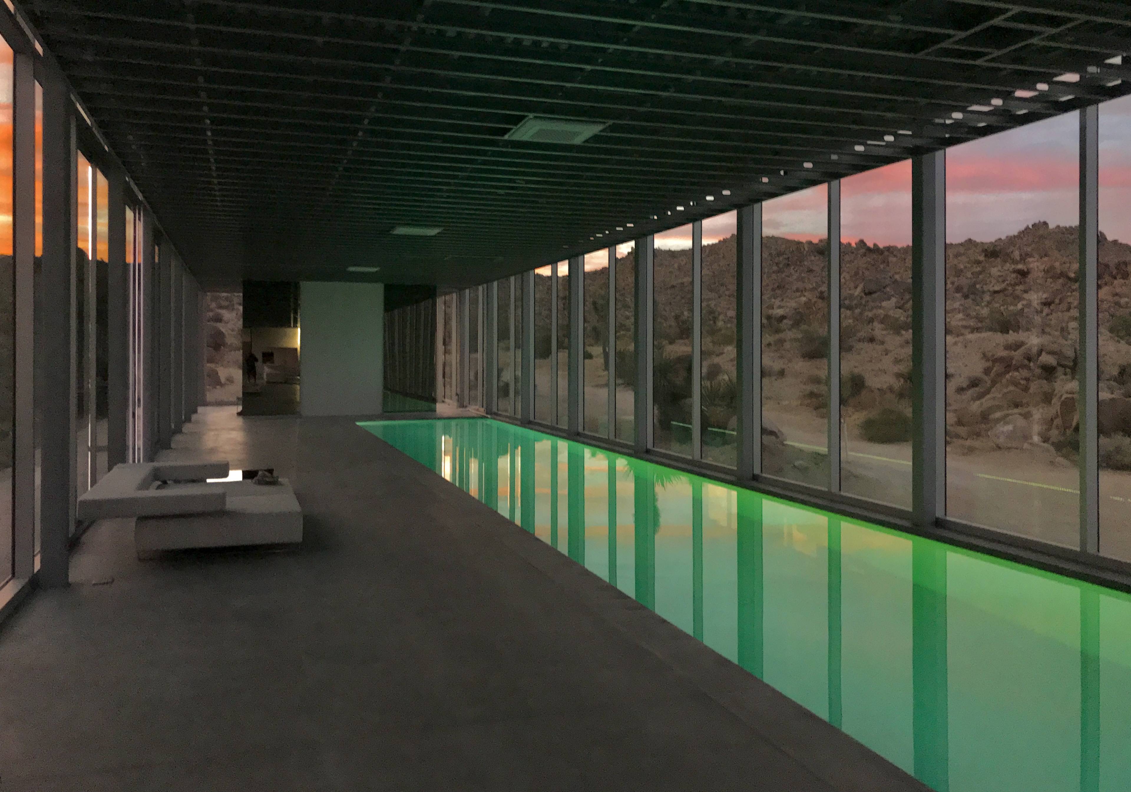 The image shows a modern, minimalist indoor pool area with large glass walls overlooking a rocky landscape at sunset.