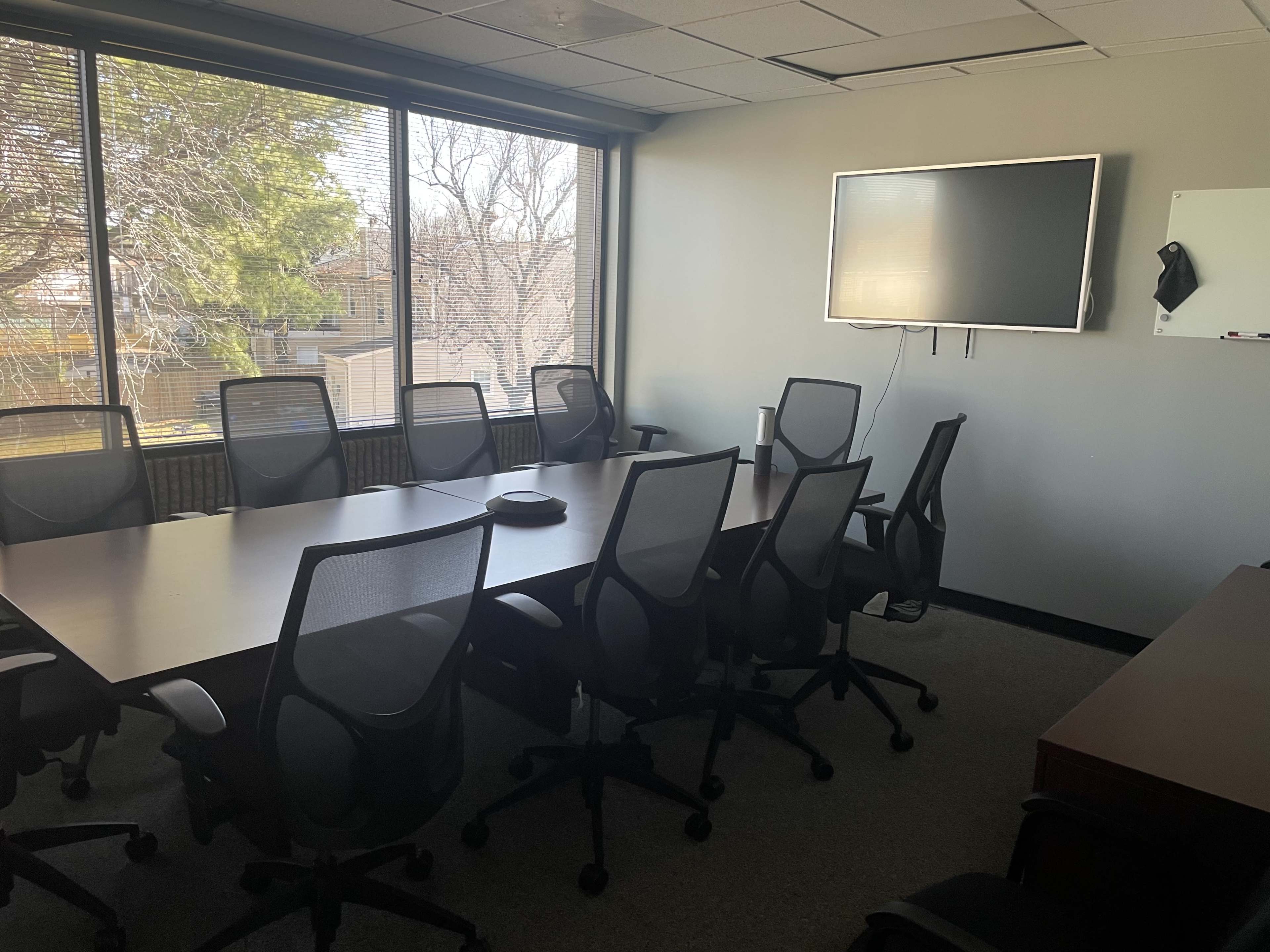 A meeting room with a large conference table surrounded by eight chairs, a wall-mounted television, and a whiteboard, all set against a backdrop of large windows.