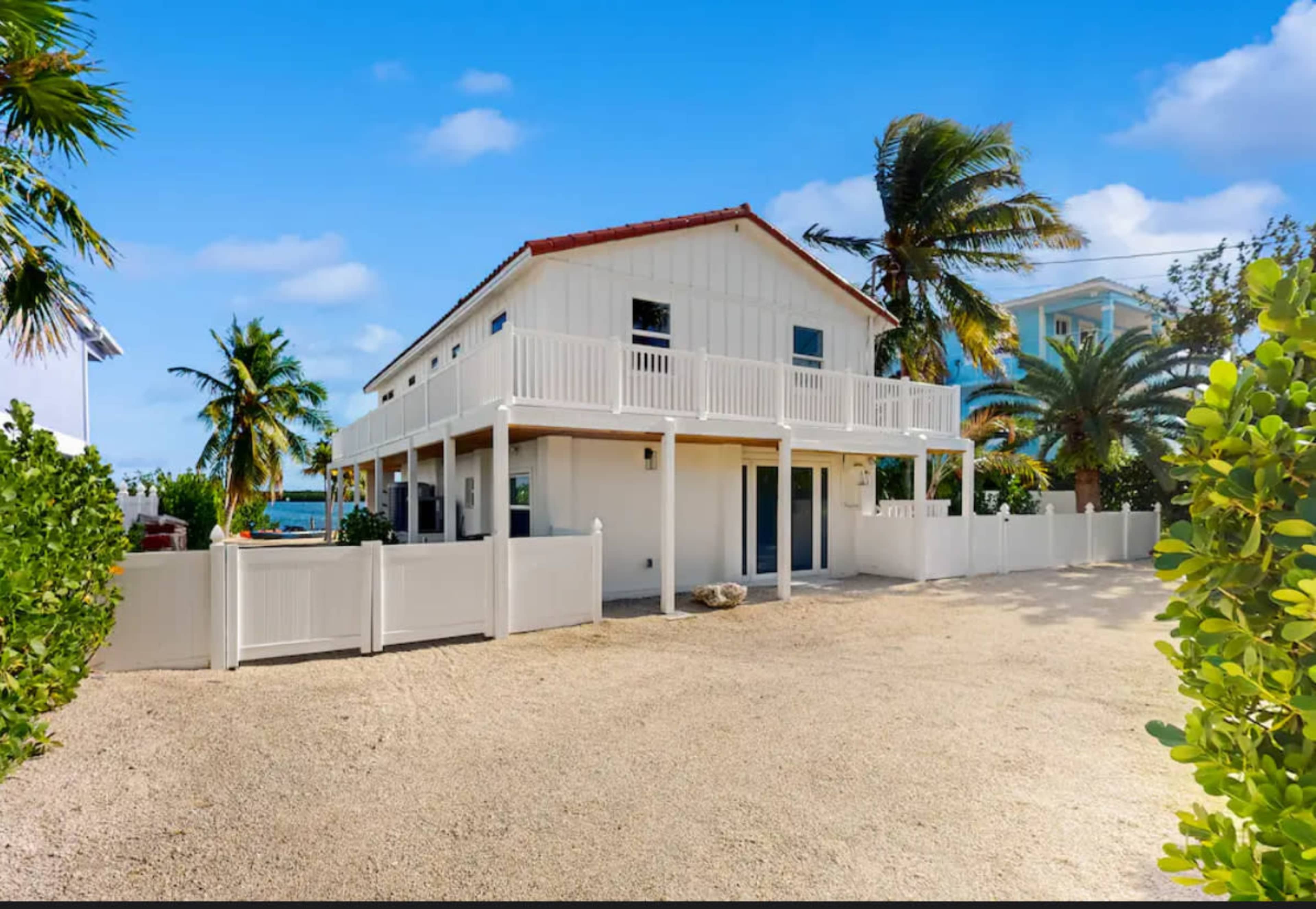 A two-story coastal house with a white exterior and a red-tiled roof is surrounded by palm trees and a white fence.