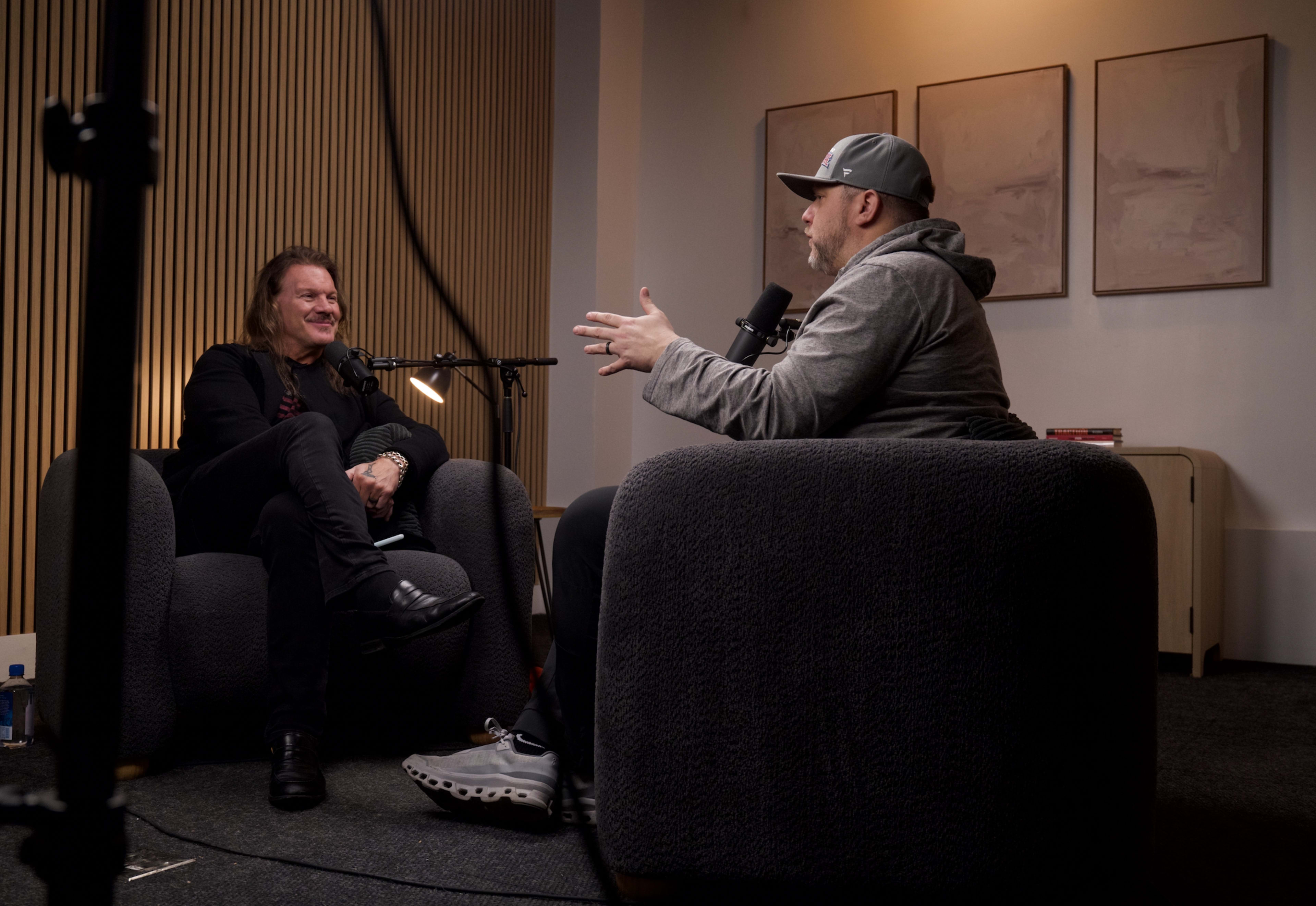 Two men are sitting in plush armchairs, engaging in a discussion while microphones are positioned in front of them in a well-lit room with minimalist decor.