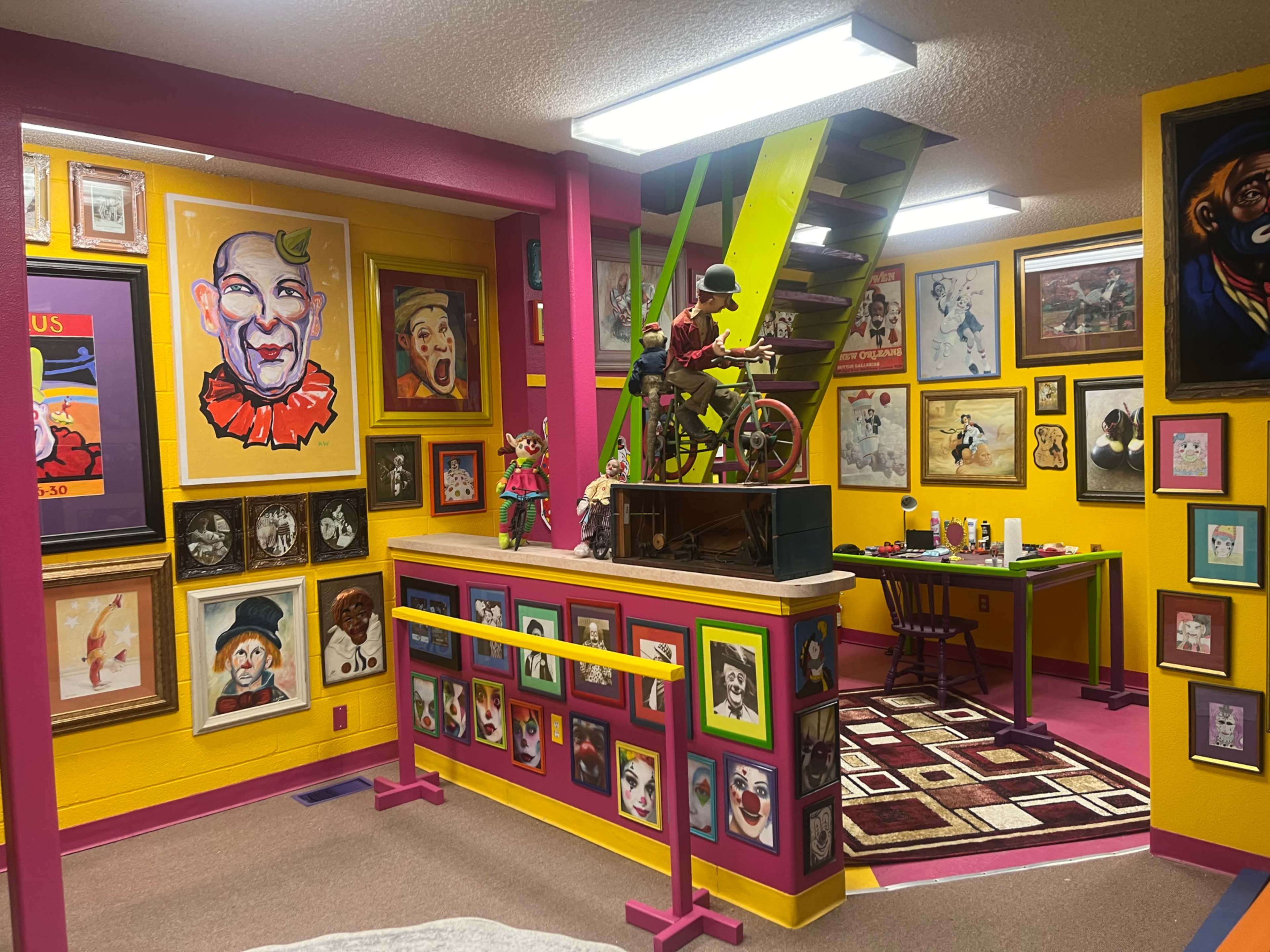 The image shows a vibrant and colorful room adorned with numerous framed clown portraits, a staircase with green railings, and a small workspace with art supplies.