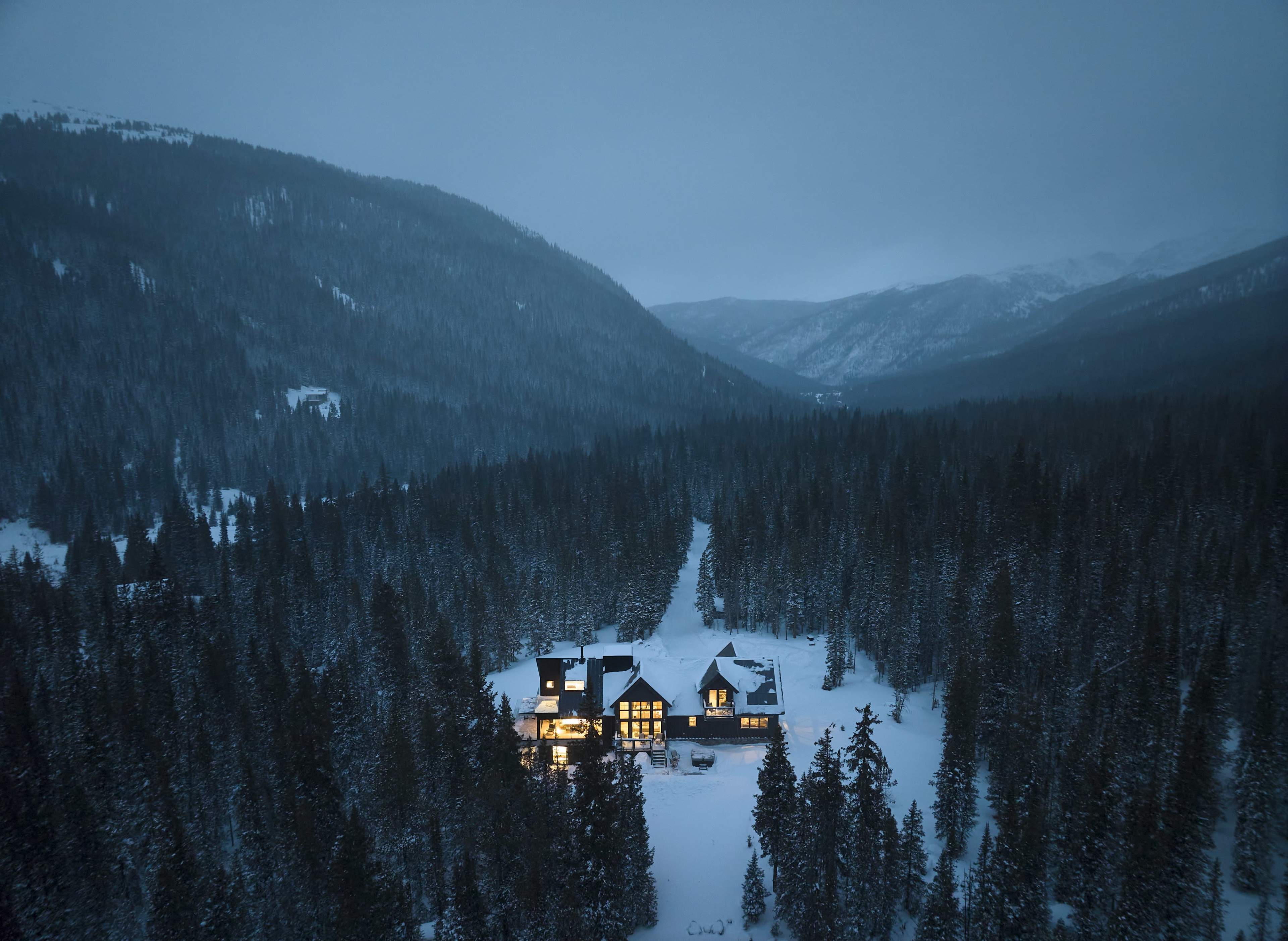 A large house sits in a snow-covered valley surrounded by evergreen trees, with dim lights illuminating its windows against the backdrop of a darkening sky.