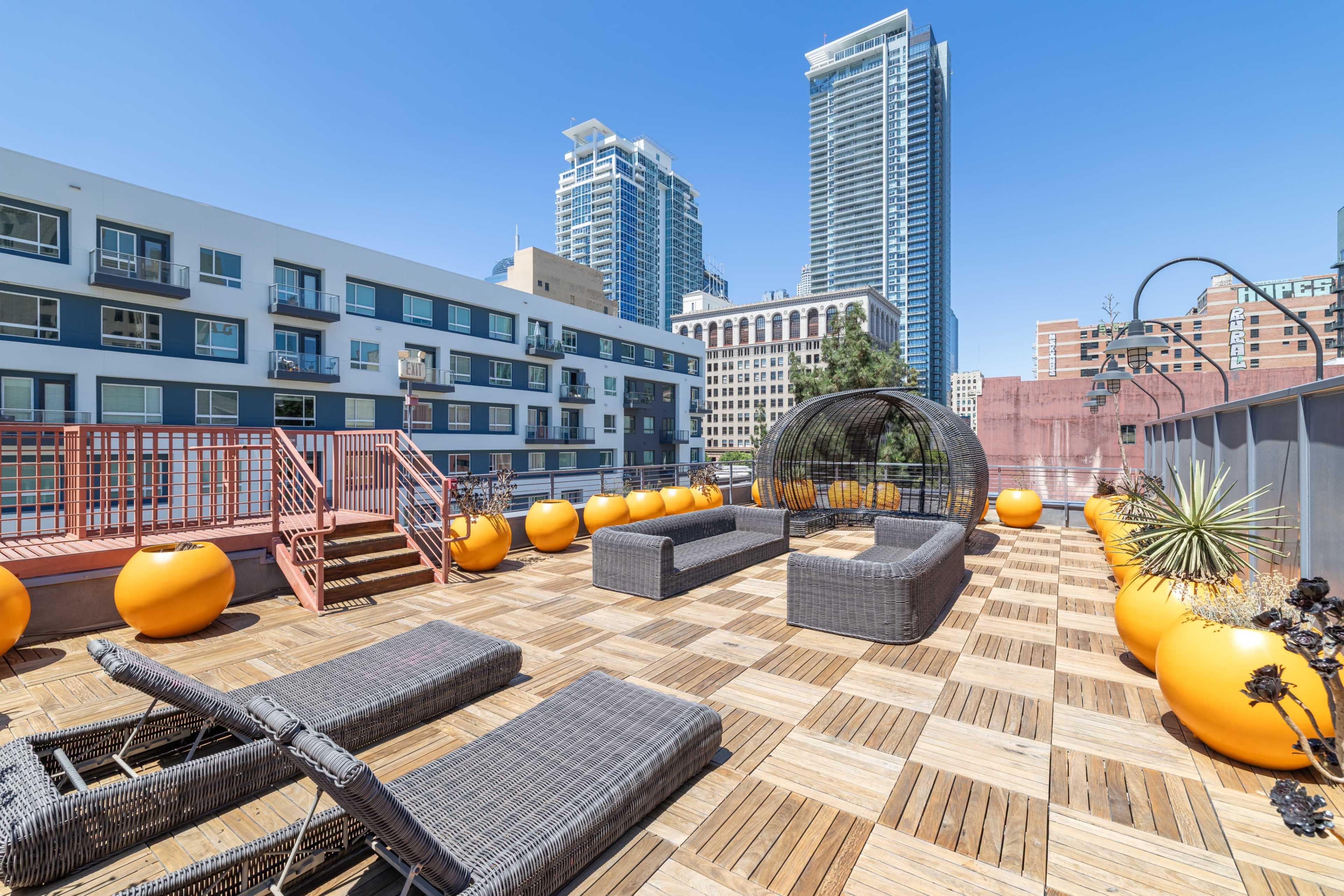 The rooftop space features a wooden deck with lounge chairs and large orange planters, surrounded by tall city buildings under a clear blue sky.