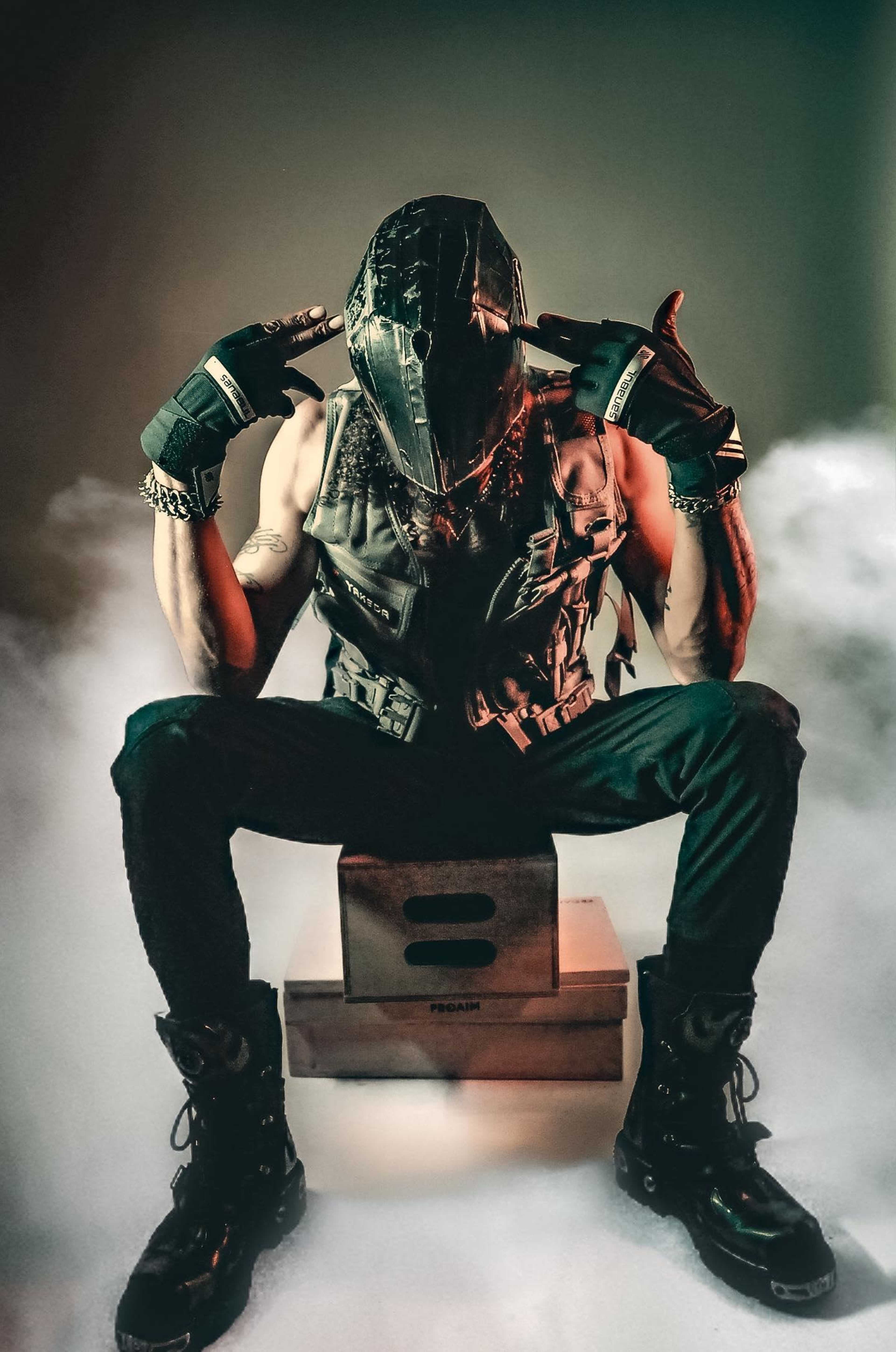 A person wearing a mask and a vest sits on a box, making a gesture with their hands amid smoke.