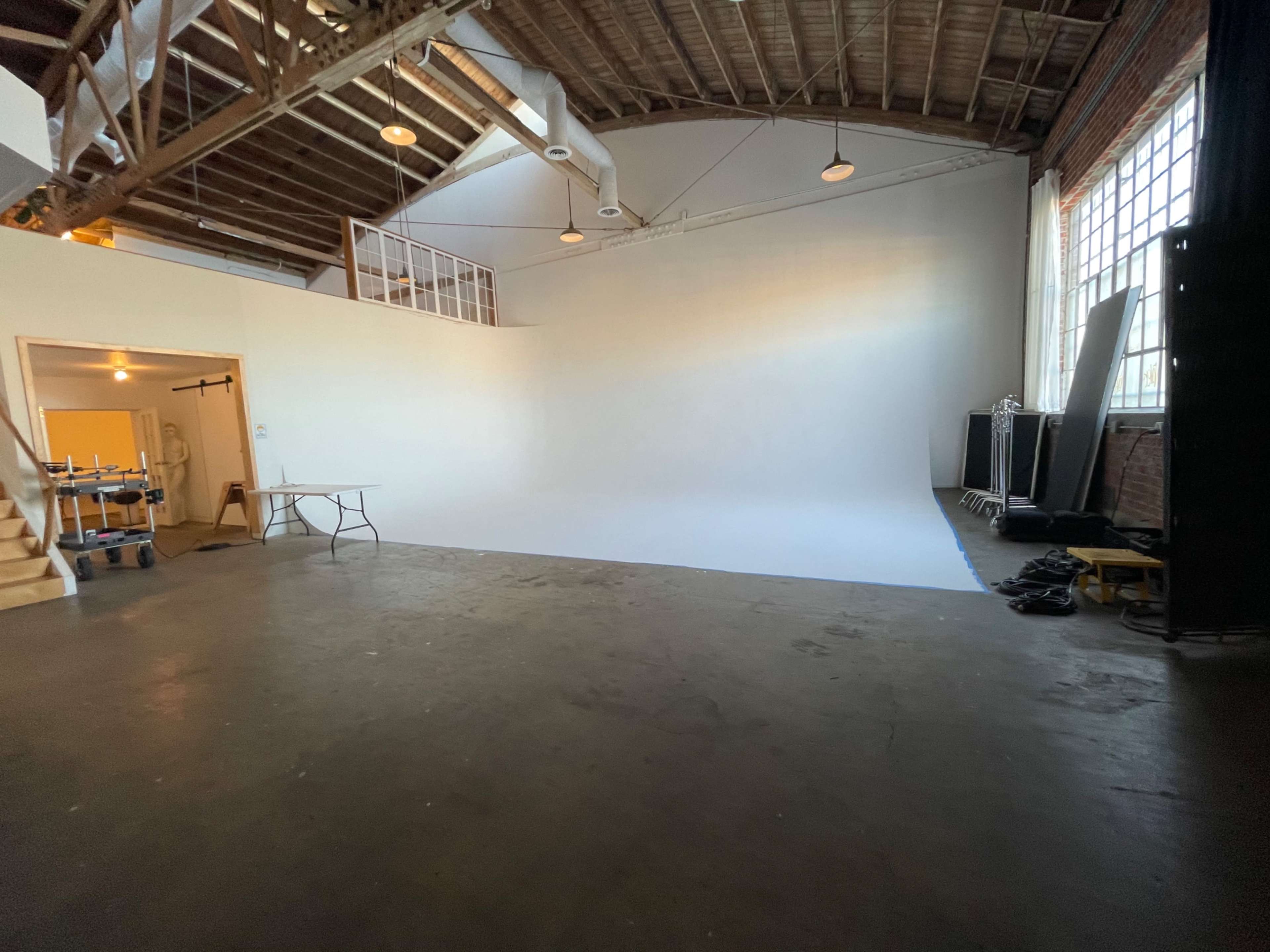 The image shows a spacious studio with a white backdrop, wooden beams, and a polished floor, along with a table and some equipment along the side.