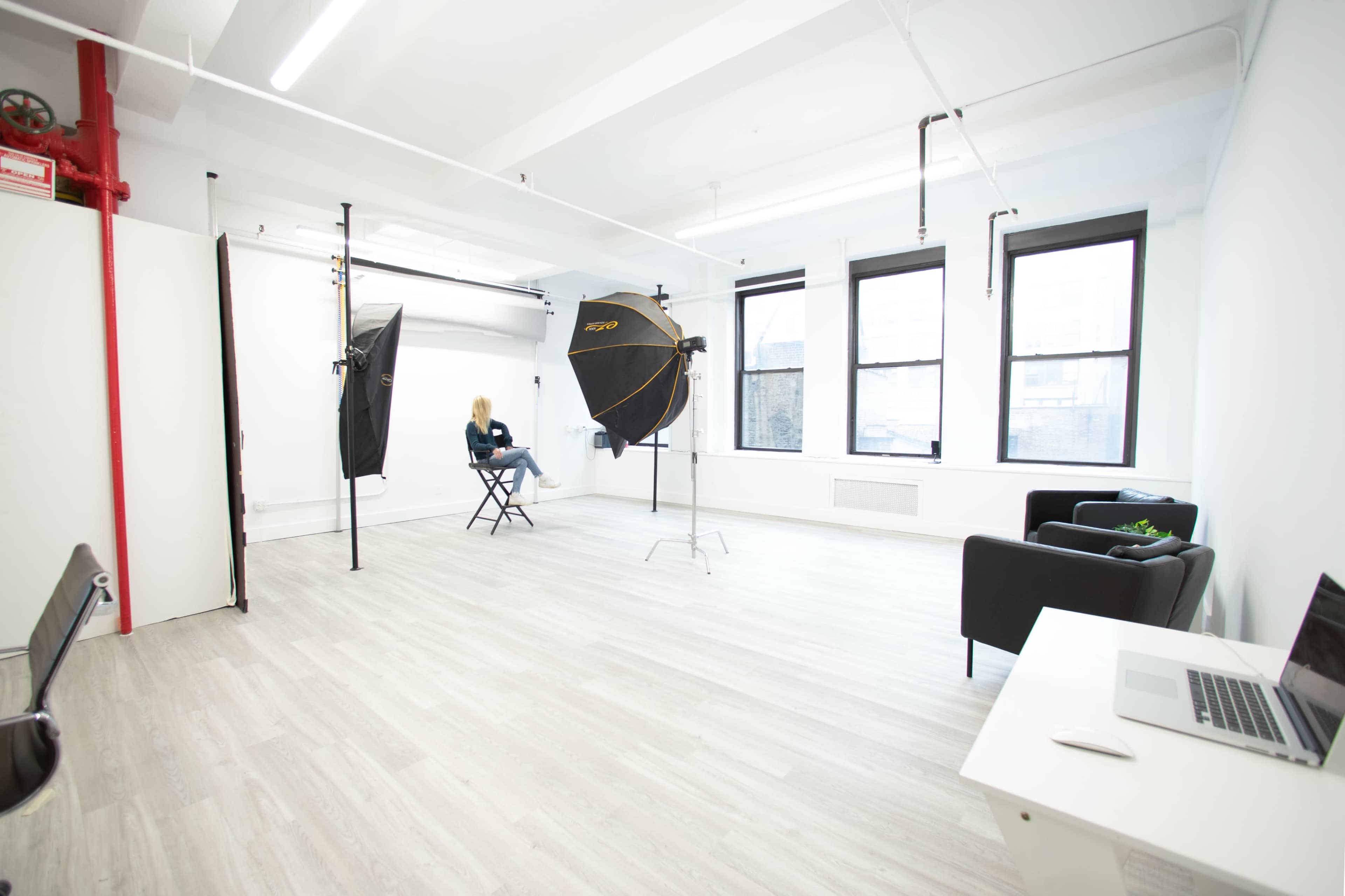 A bright photography studio with two softbox lights, a vacant chair, and a small seating area.