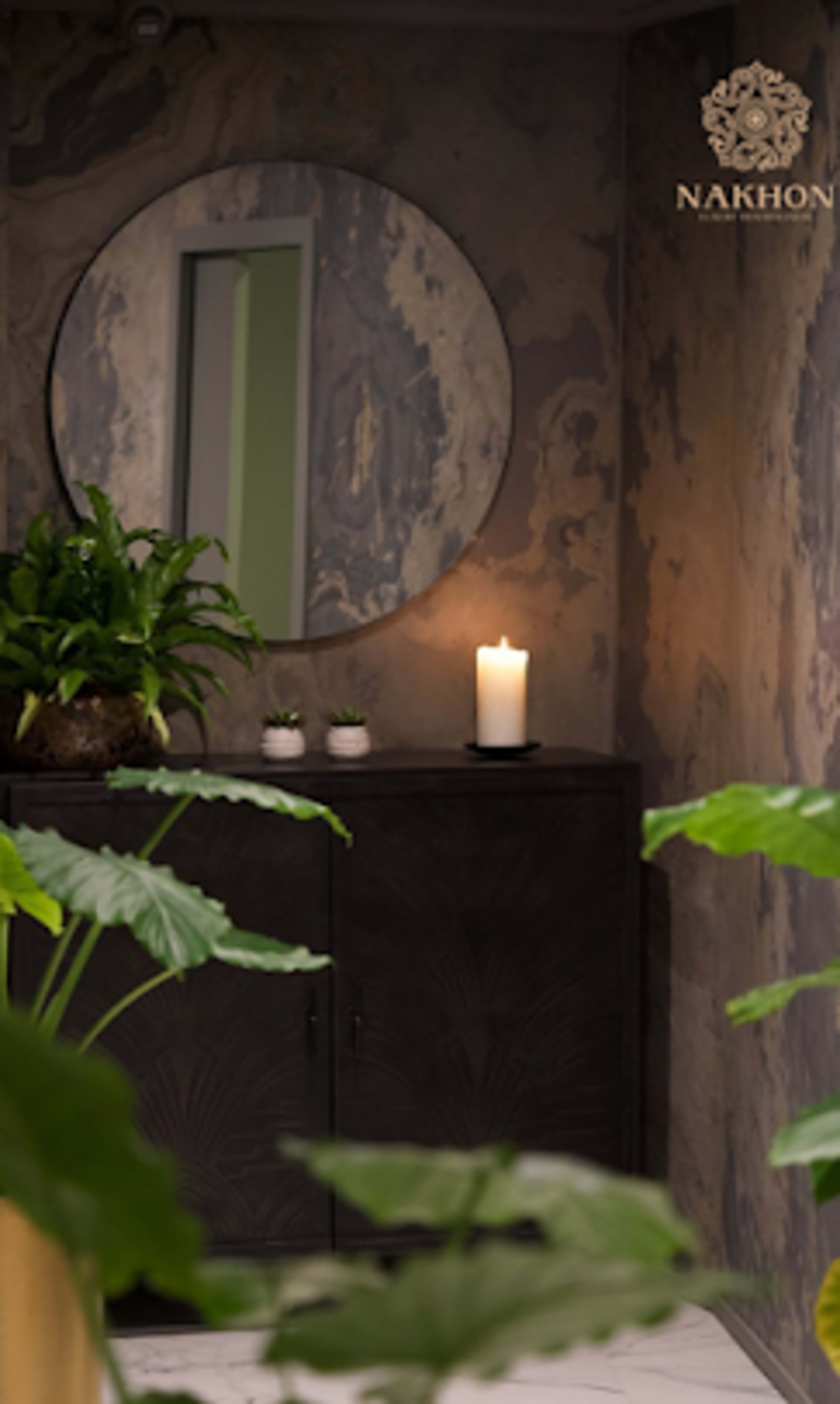The image shows a modern room with a circular mirror on the wall, a candle on a dark cabinet, and green plants in the foreground.