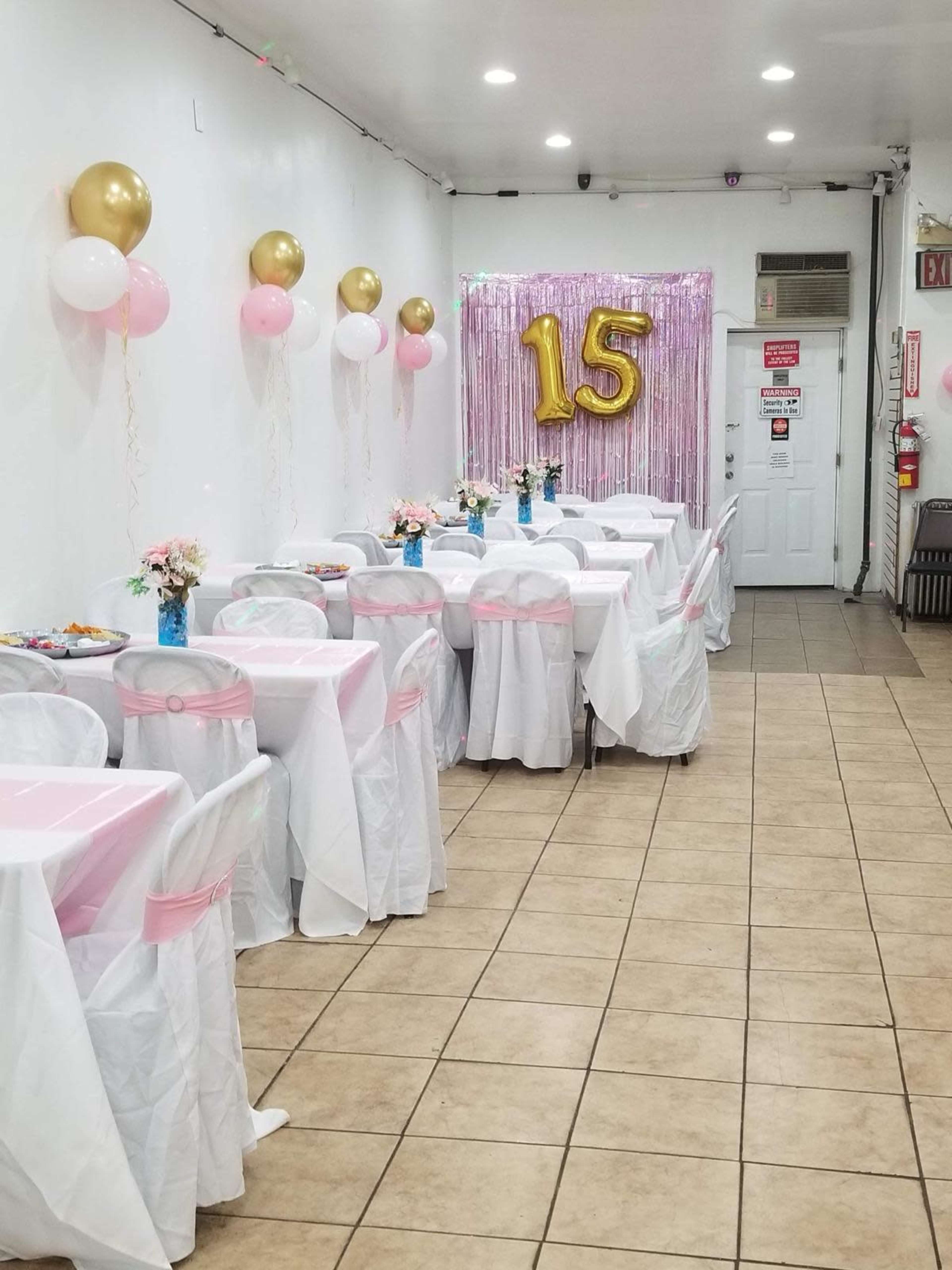 The room is set up for a celebration with tables dressed in white tablecloths and pink accents, adorned with flowers, balloons, and a large golden "15" decoration on the wall.