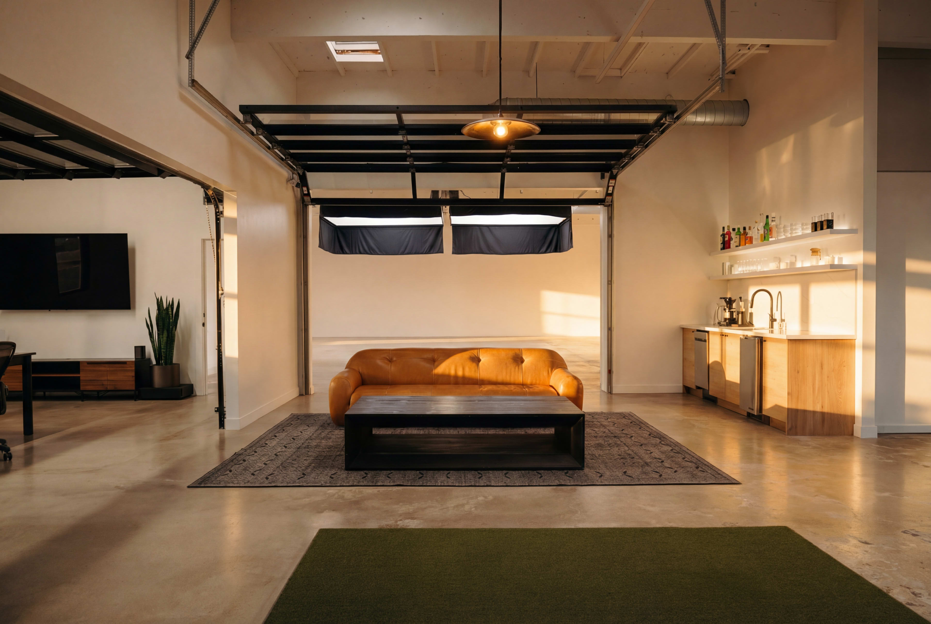 The image shows a modern, spacious living area with a tan leather sofa, a dark coffee table, a bar area, and large garage-style windows letting in sunlight.