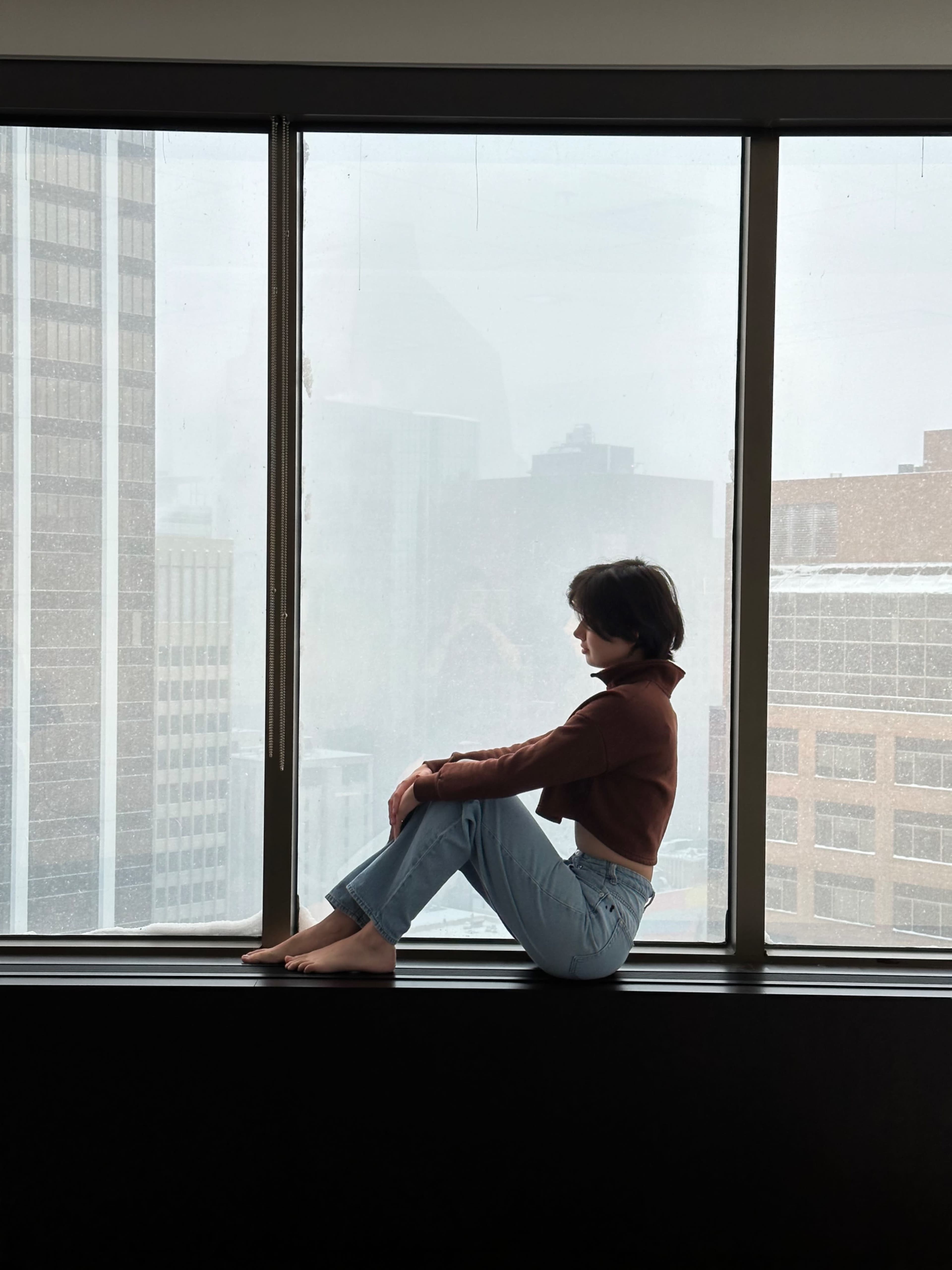 A person sits cross-legged on a windowsill, gazing out at a cityscape obscured by fog.