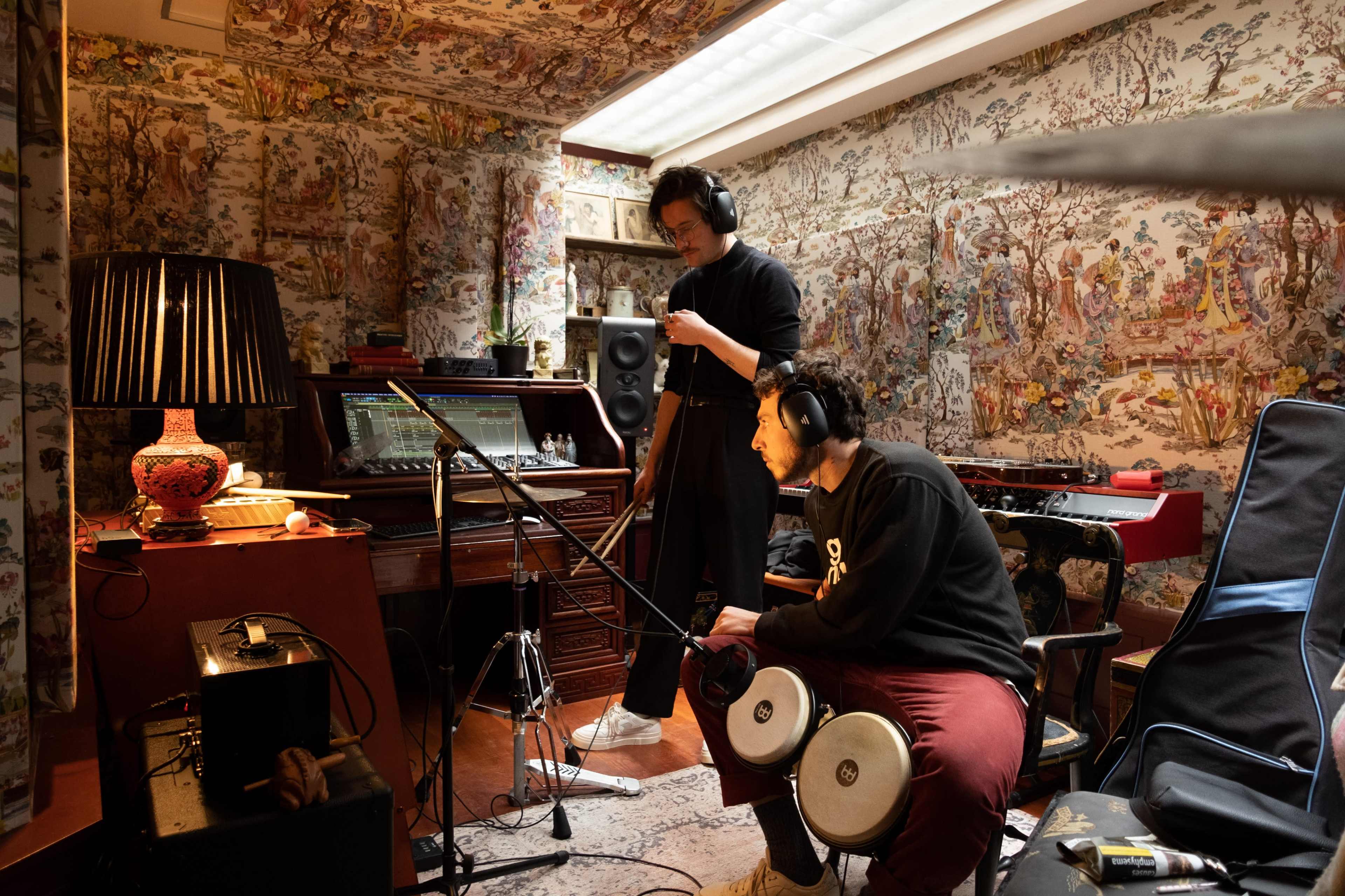 Two individuals wearing headphones are working in a music studio decorated with patterned wallpaper, with recording equipment and instruments visible around them.