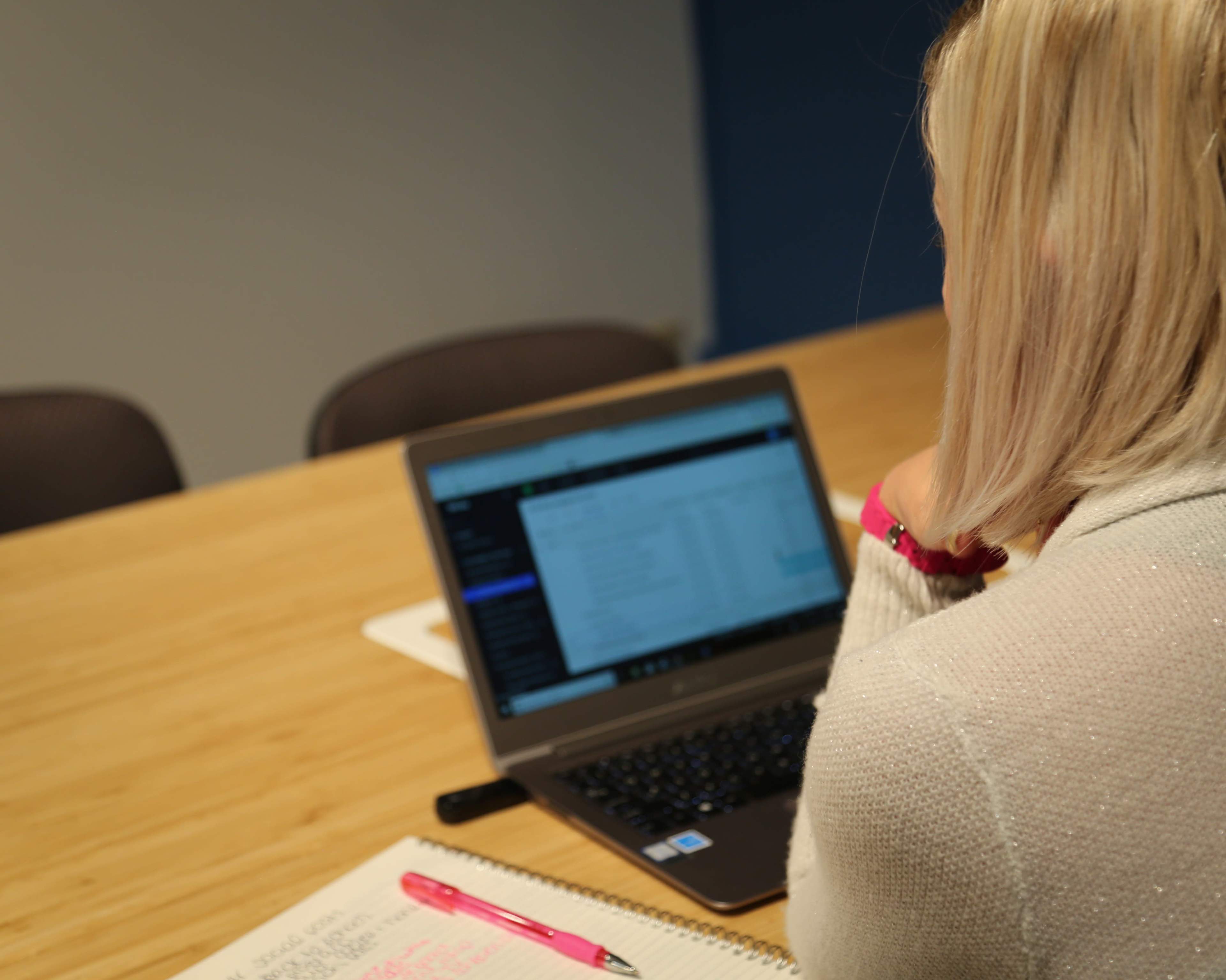 A person is sitting at a wooden table, using a laptop with a notebook and a pink pen beside them.