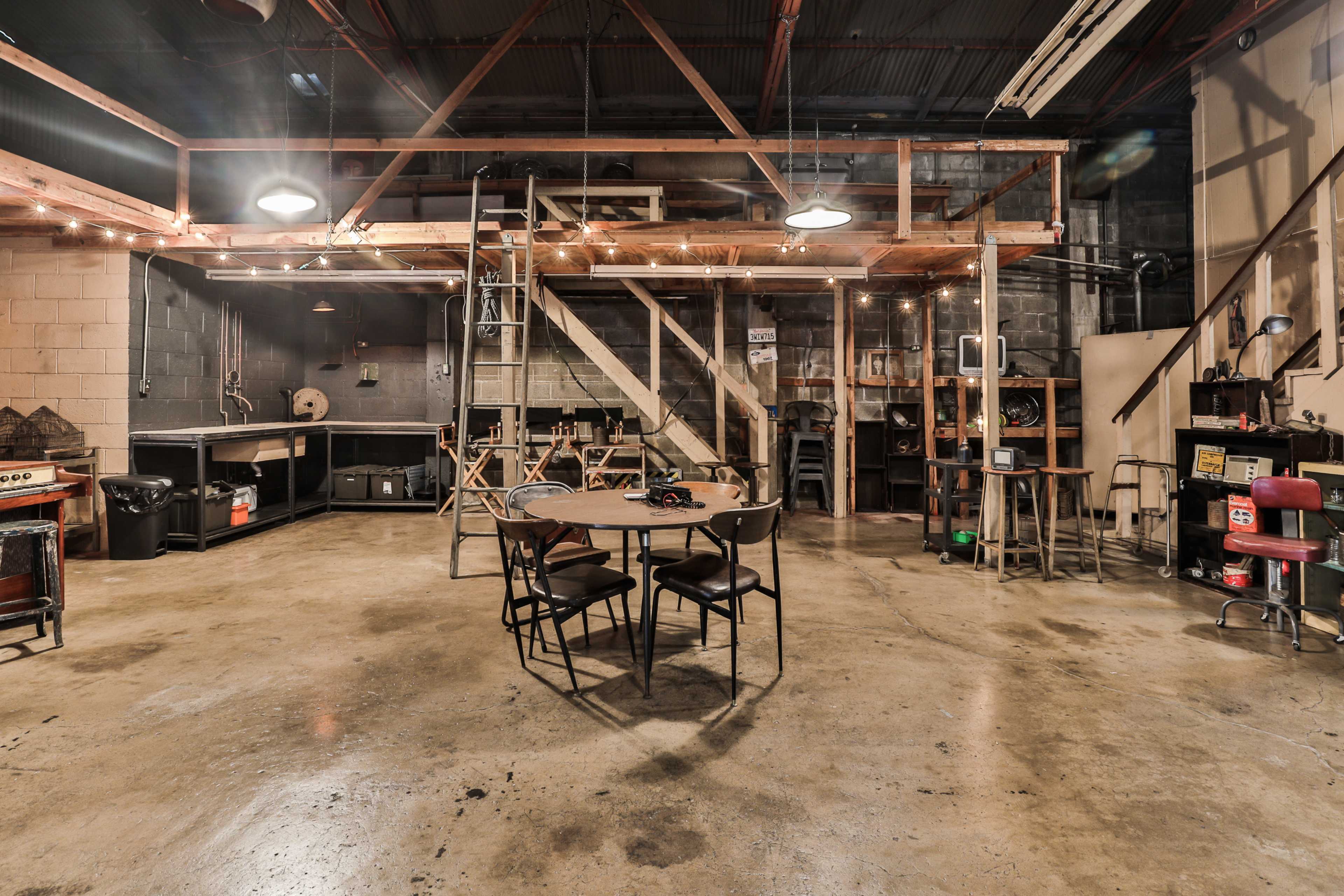 An industrial-style workshop featuring a wooden loft, a central table with chairs, and various tools and equipment along the walls.