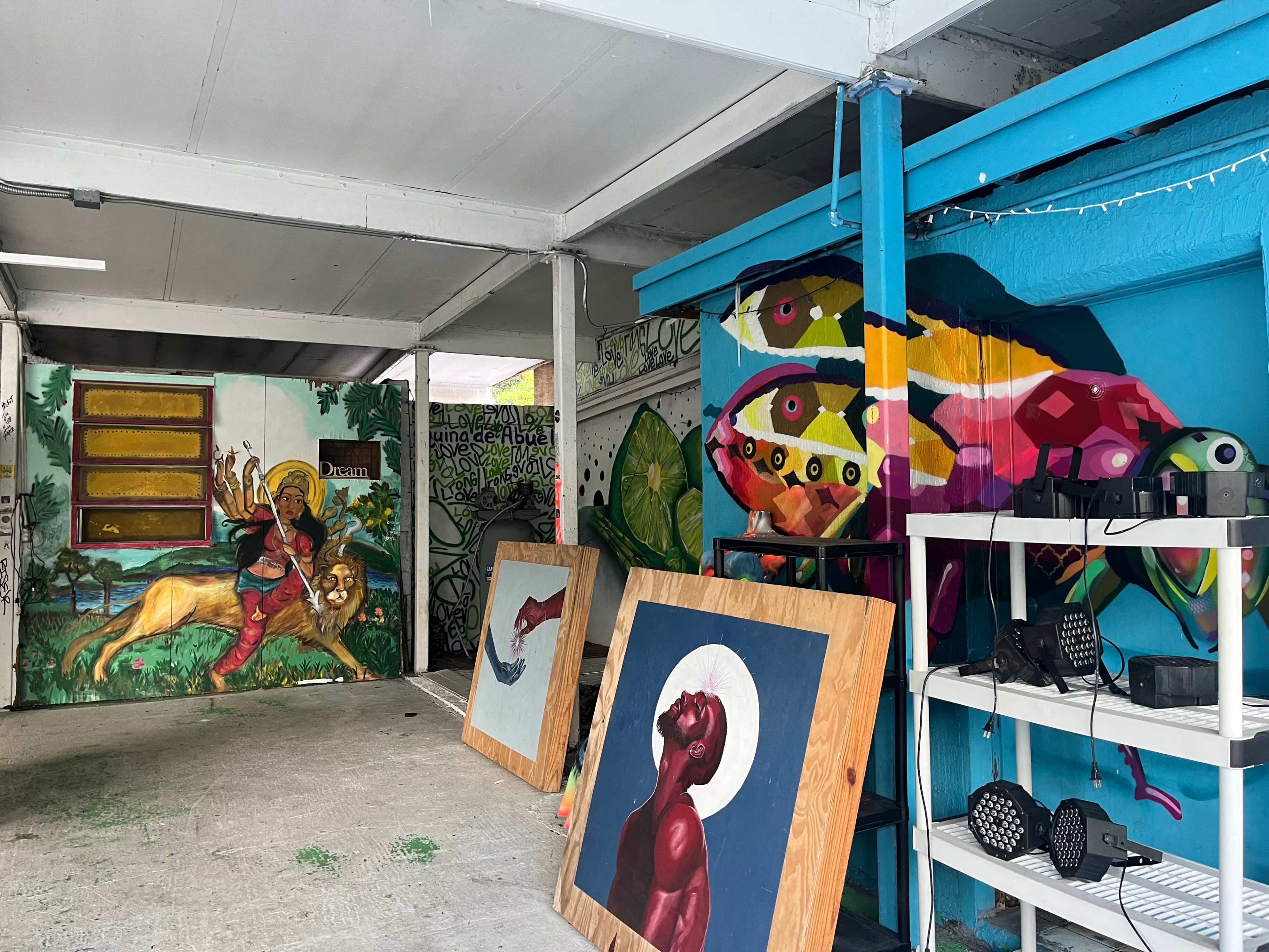 The scene shows an indoor area featuring colorful murals on the walls, with two large paintings propped against the floor and shelves holding lighting equipment.