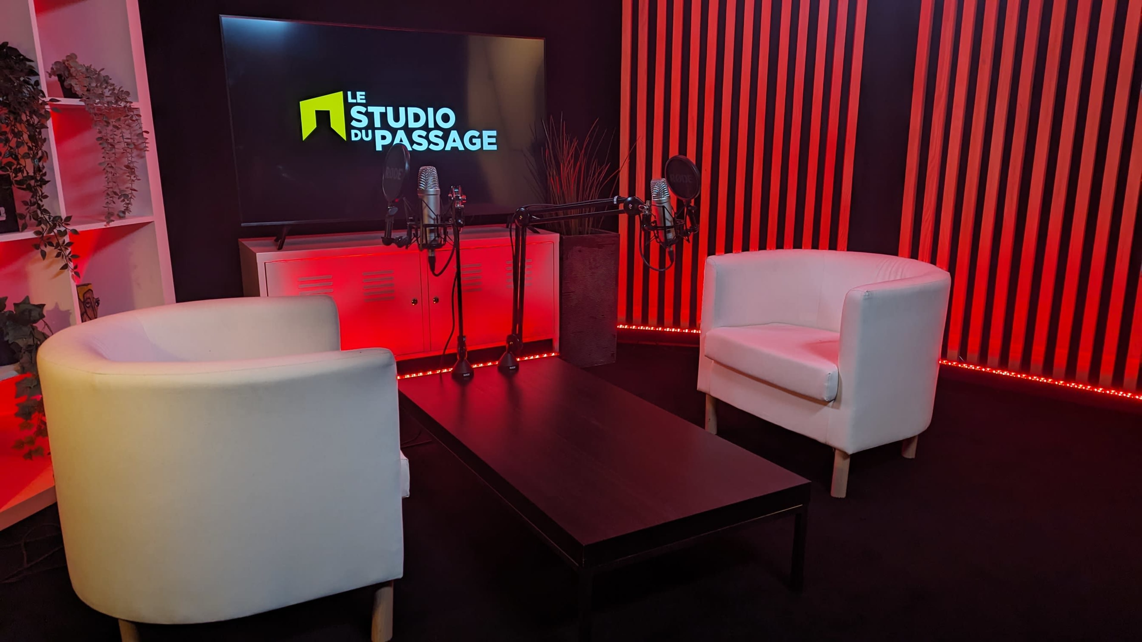 A modern studio setup with two white chairs, a black table, and two microphones in front of a screen displaying "LE STUDIO DU PASSAGE" against a backdrop of red and black stripes.