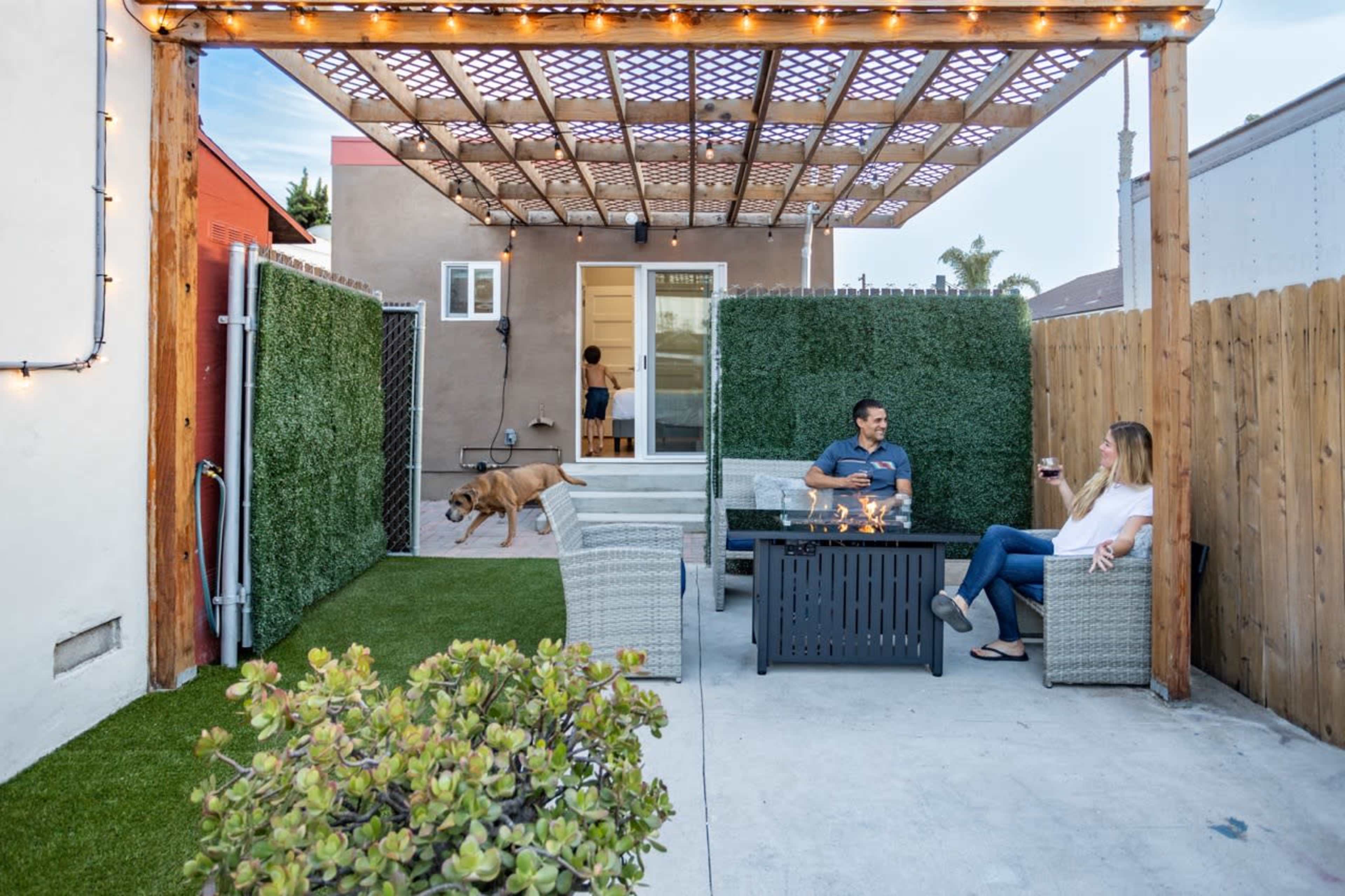 A man and a woman sit on patio furniture under a pergola with string lights, while a dog runs nearby in a small outdoor space.