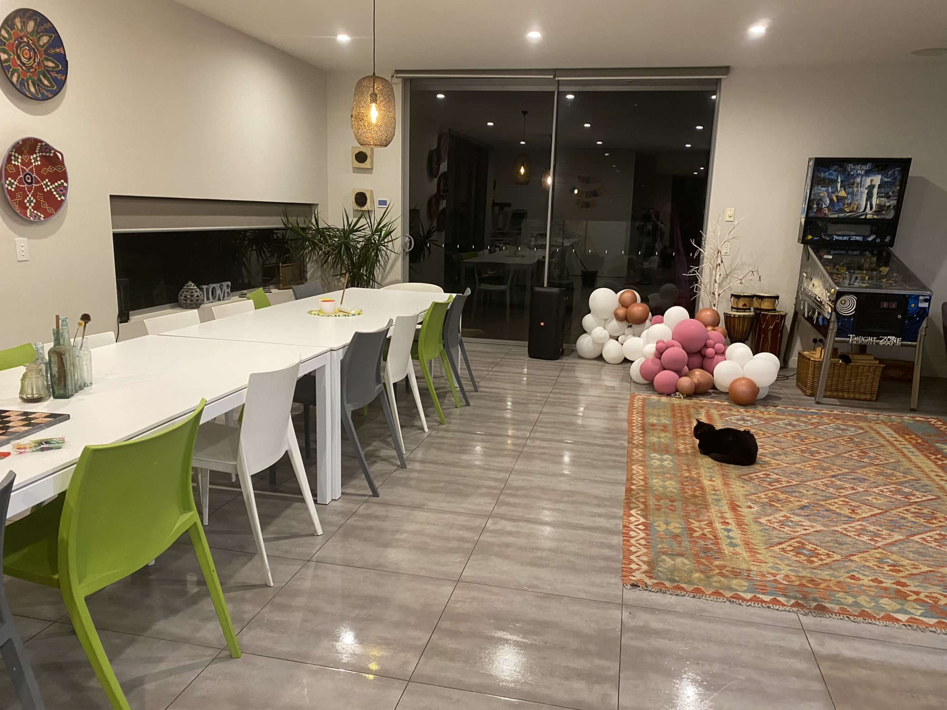 A spacious dining area features a long white table with colorful chairs, a patterned rug on the floor, and a black cat resting nearby.