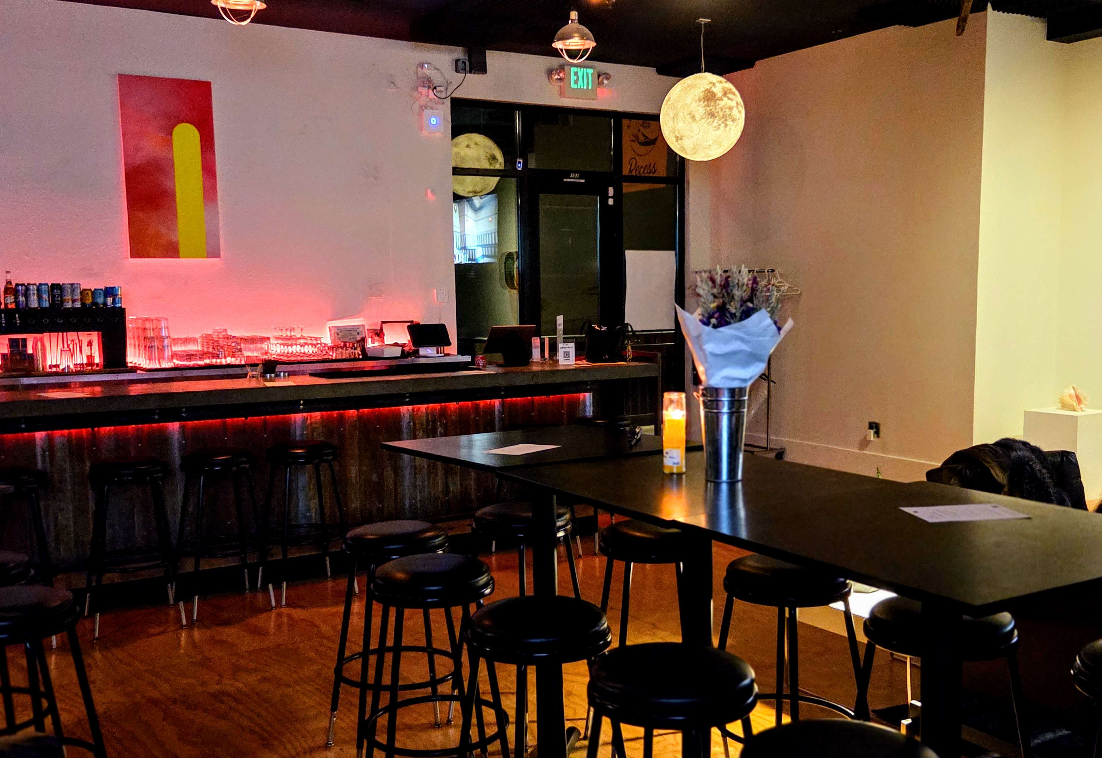 The image shows a modern bar interior featuring a long bar counter with stools, a central table, and ambient lighting.