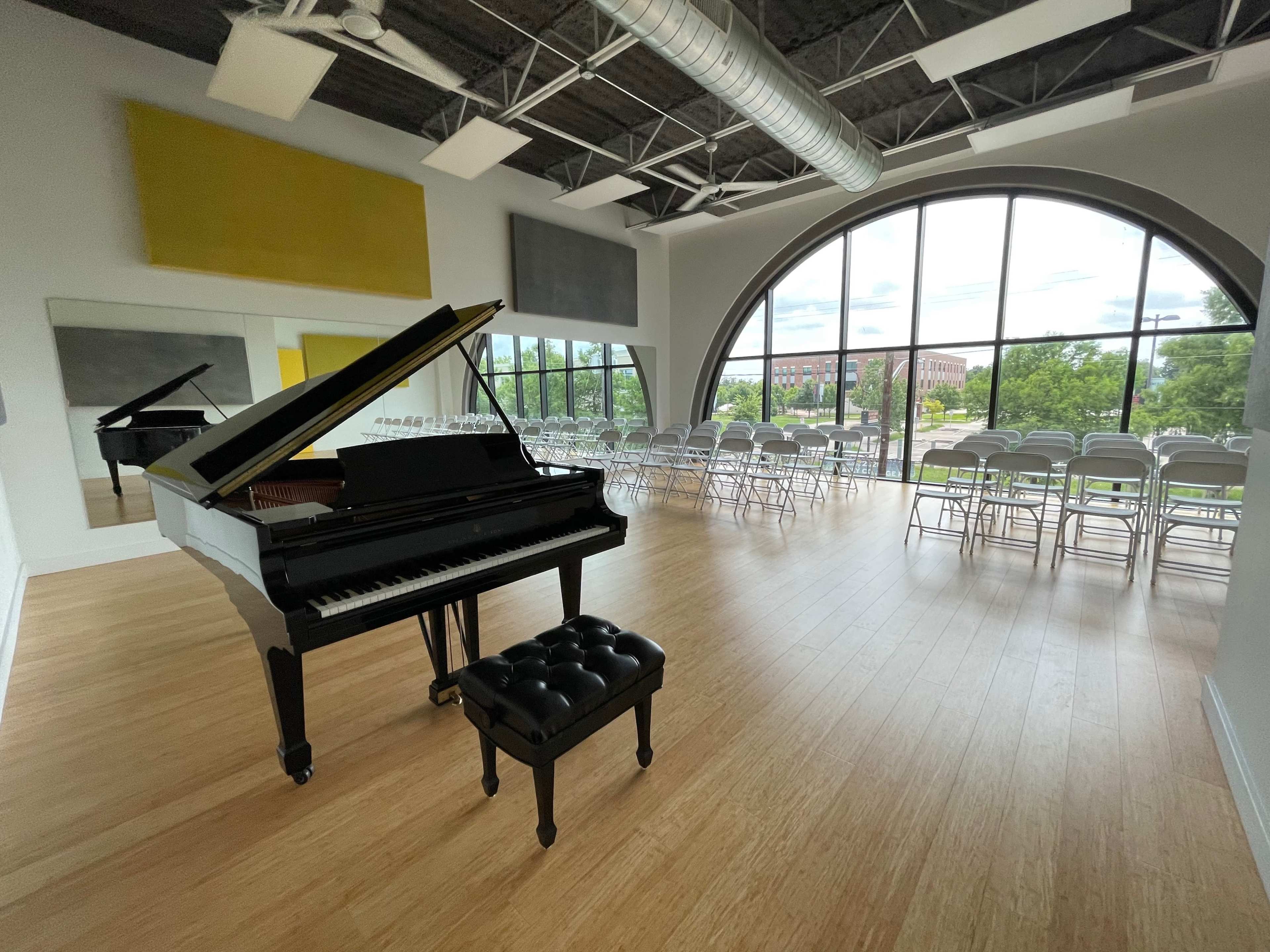A grand piano is positioned near a large window in a spacious room filled with folding chairs set up for an event.