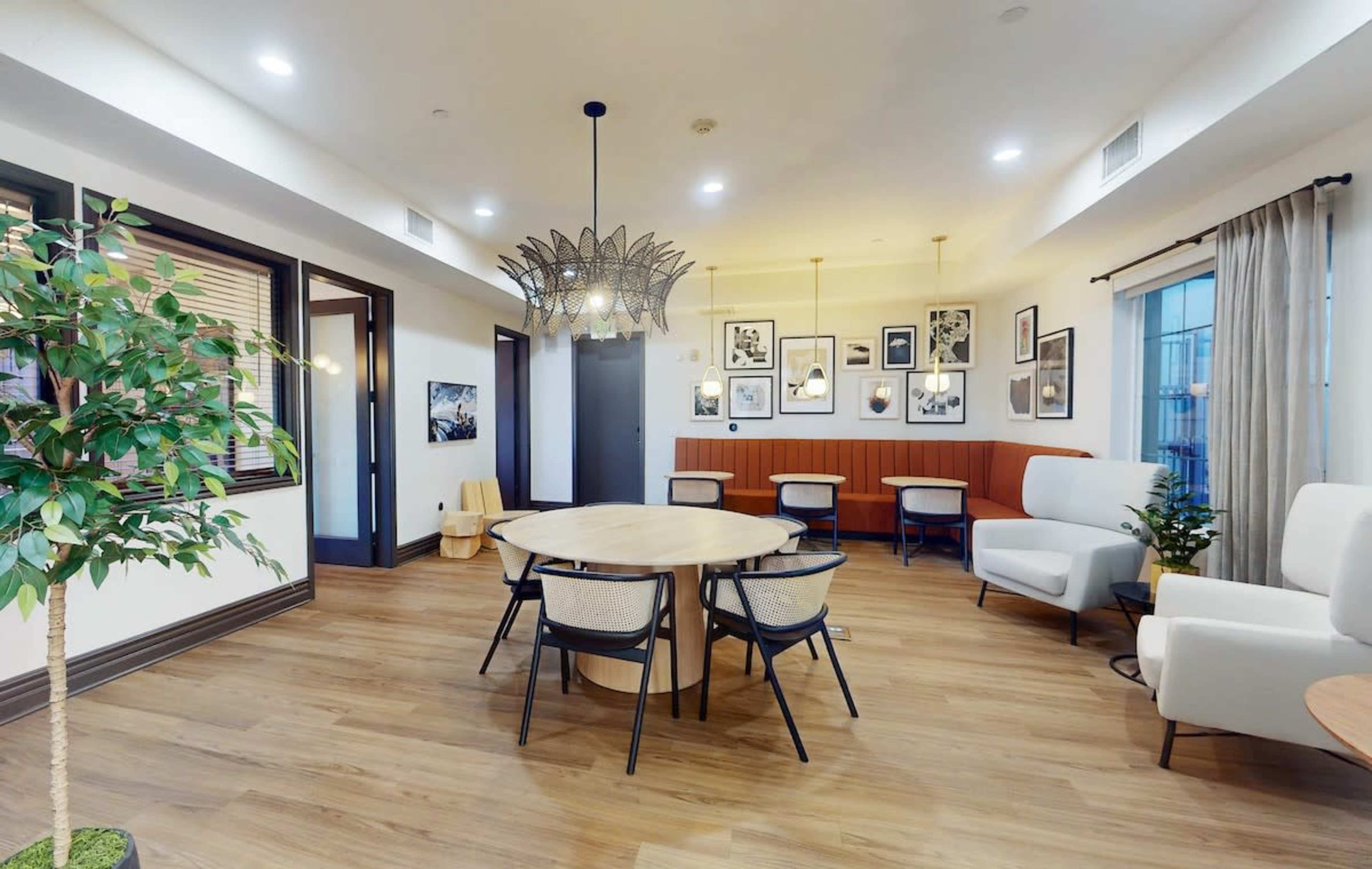 The image shows a modern lounge area featuring a round table surrounded by chairs, decorative plants, and various framed artworks on the walls.