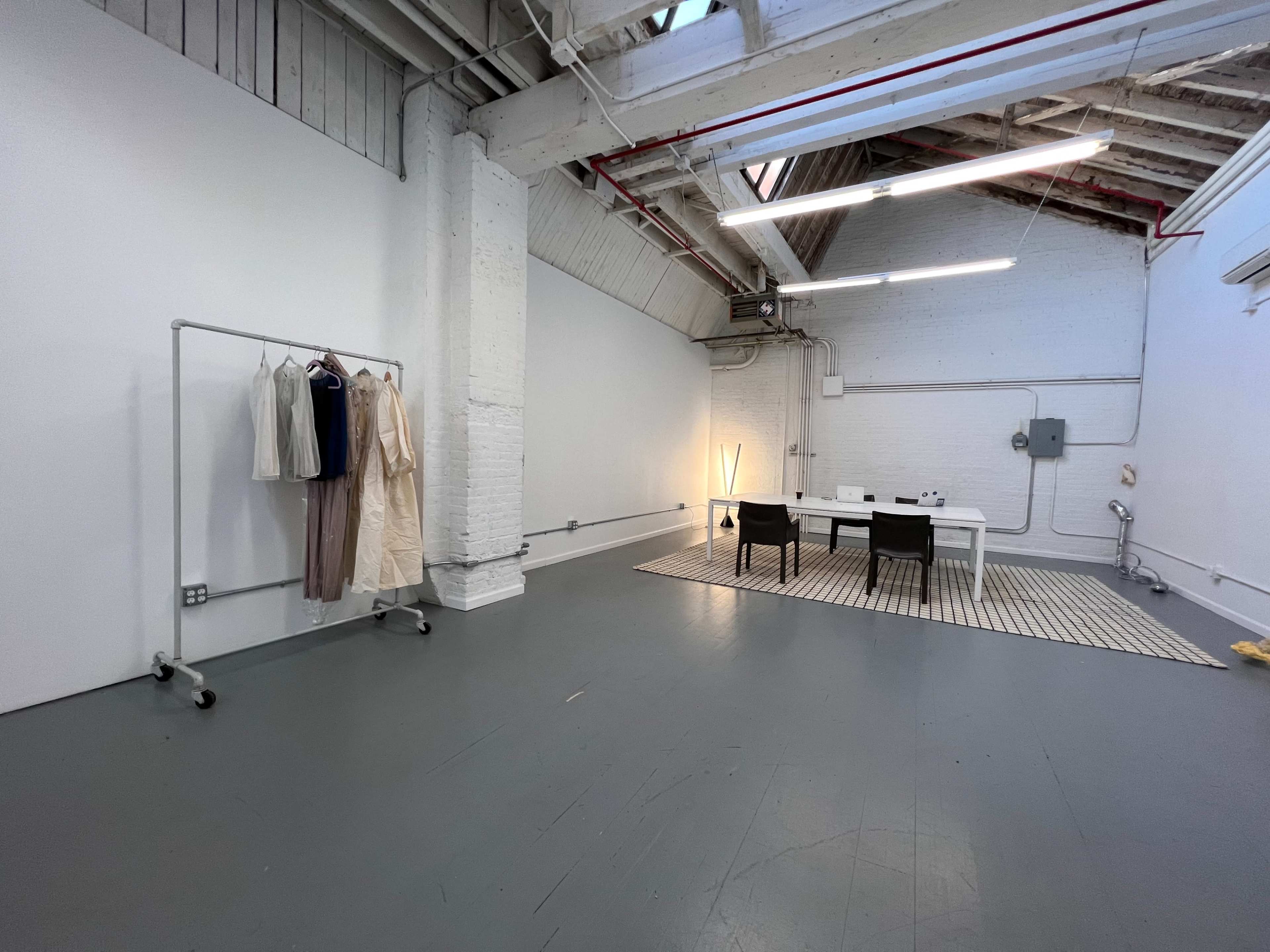 A large, open studio space with a clothing rack on one side, a table with chairs in the center, and a patterned rug beneath it.