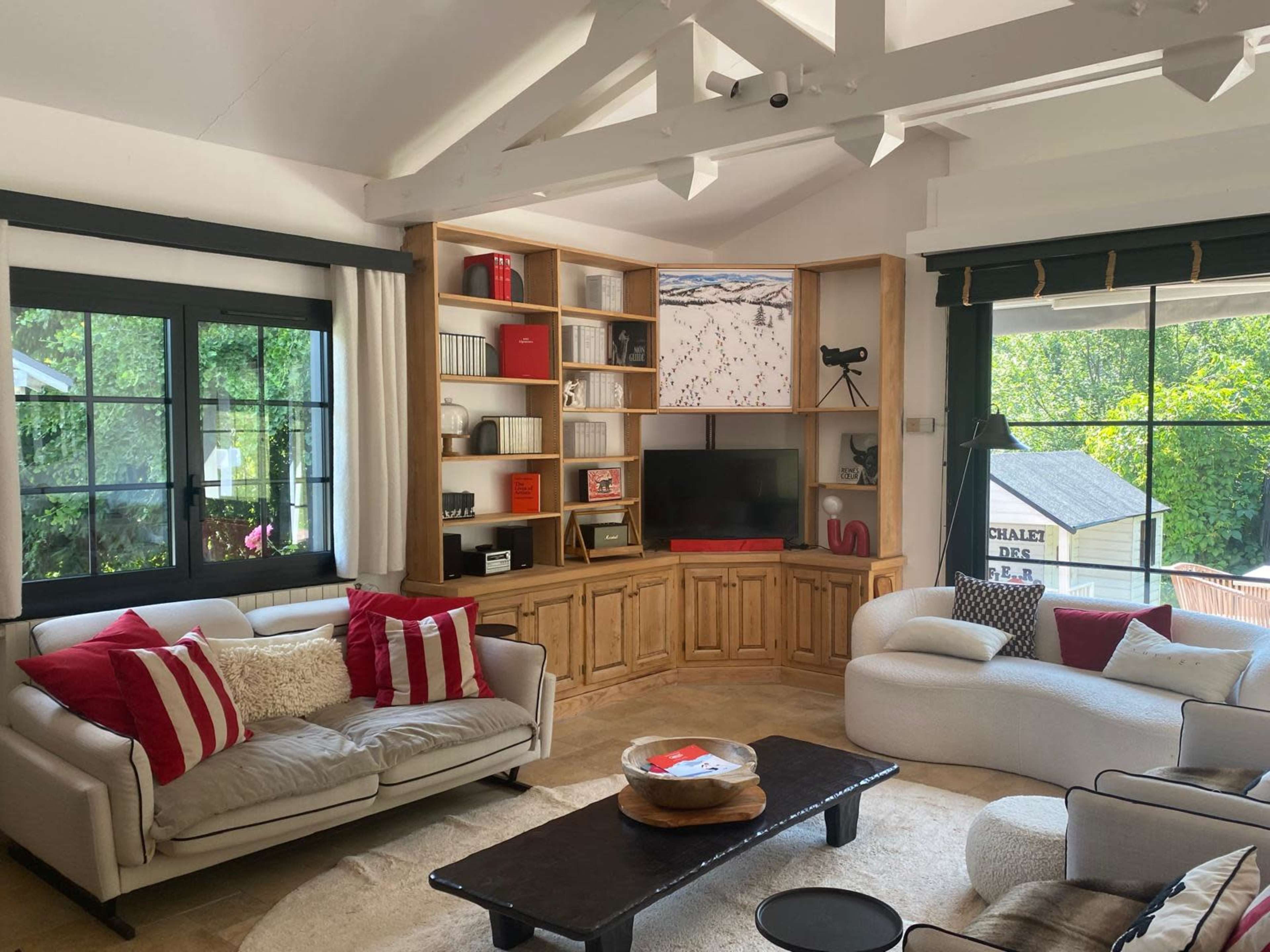The image shows a bright living room featuring a wooden entertainment unit, two sofas with cushions, and a coffee table, with large windows revealing greenery outside.