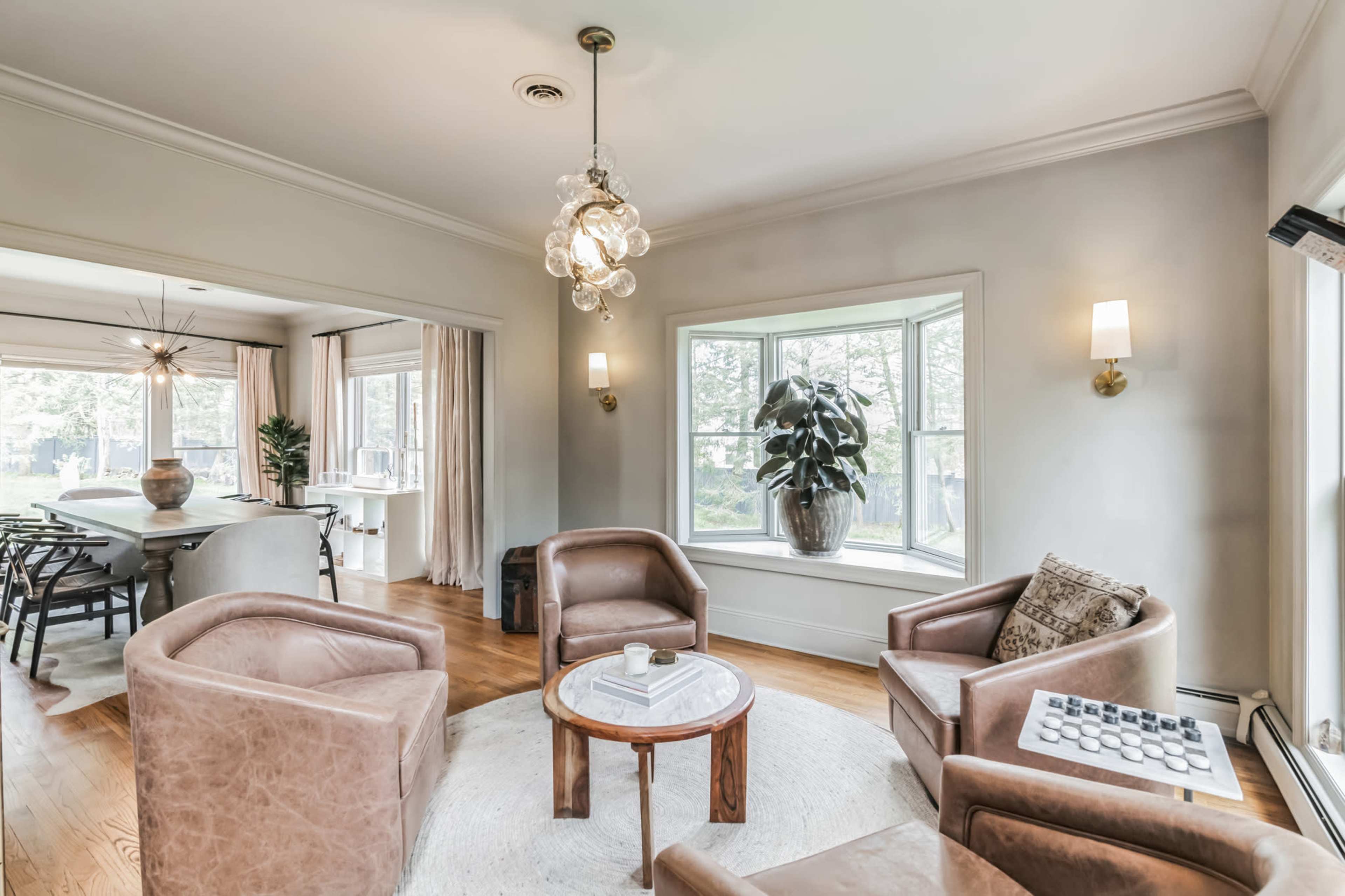 A cozy living room features four brown leather chairs arranged around a small wooden table on a circular rug, with large windows letting in natural light.