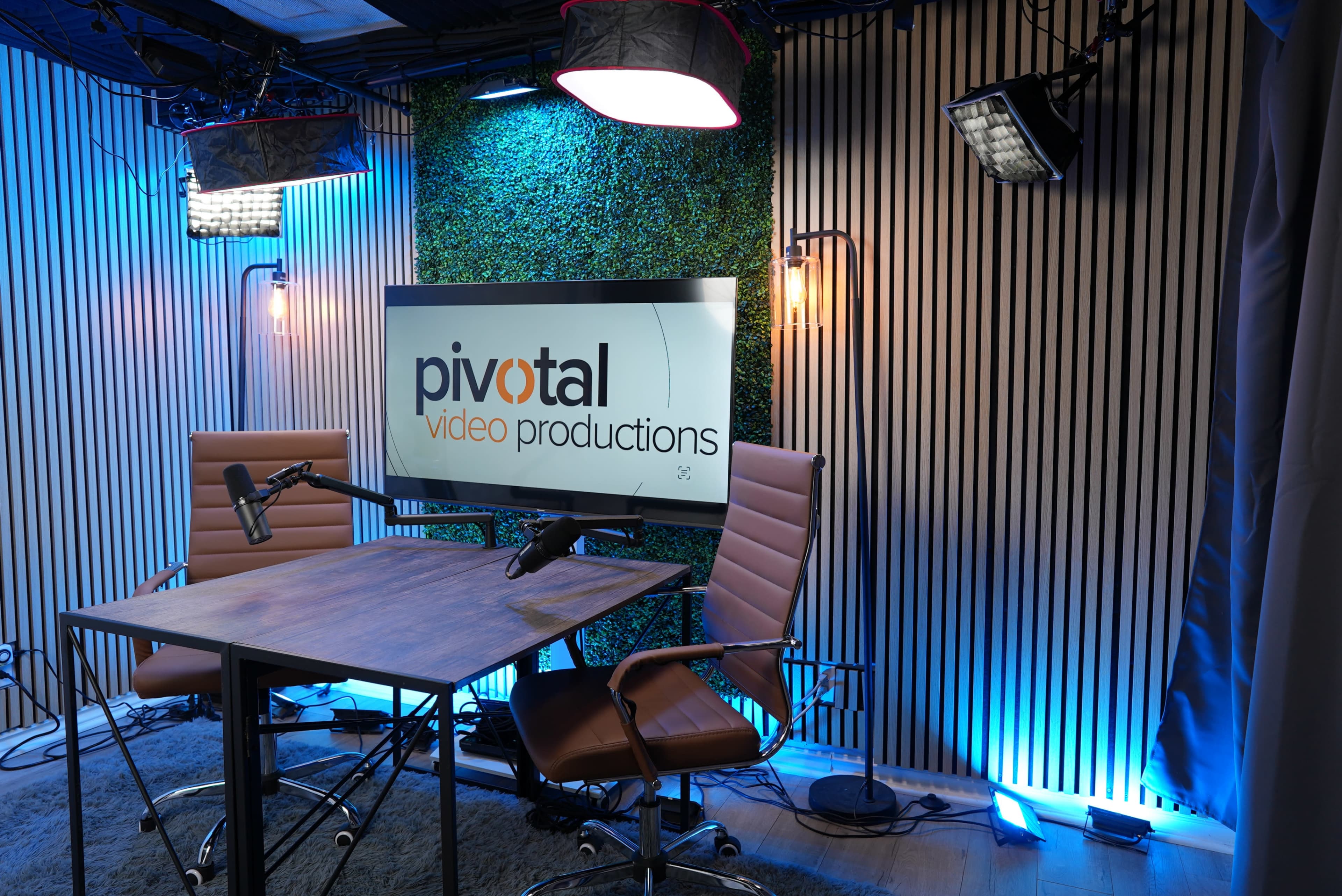 The image shows a modern video production studio setup with two chairs, a wooden table, and a large screen displaying the logo "pivotal video productions" on a backdrop of vertical wood paneling and greenery.