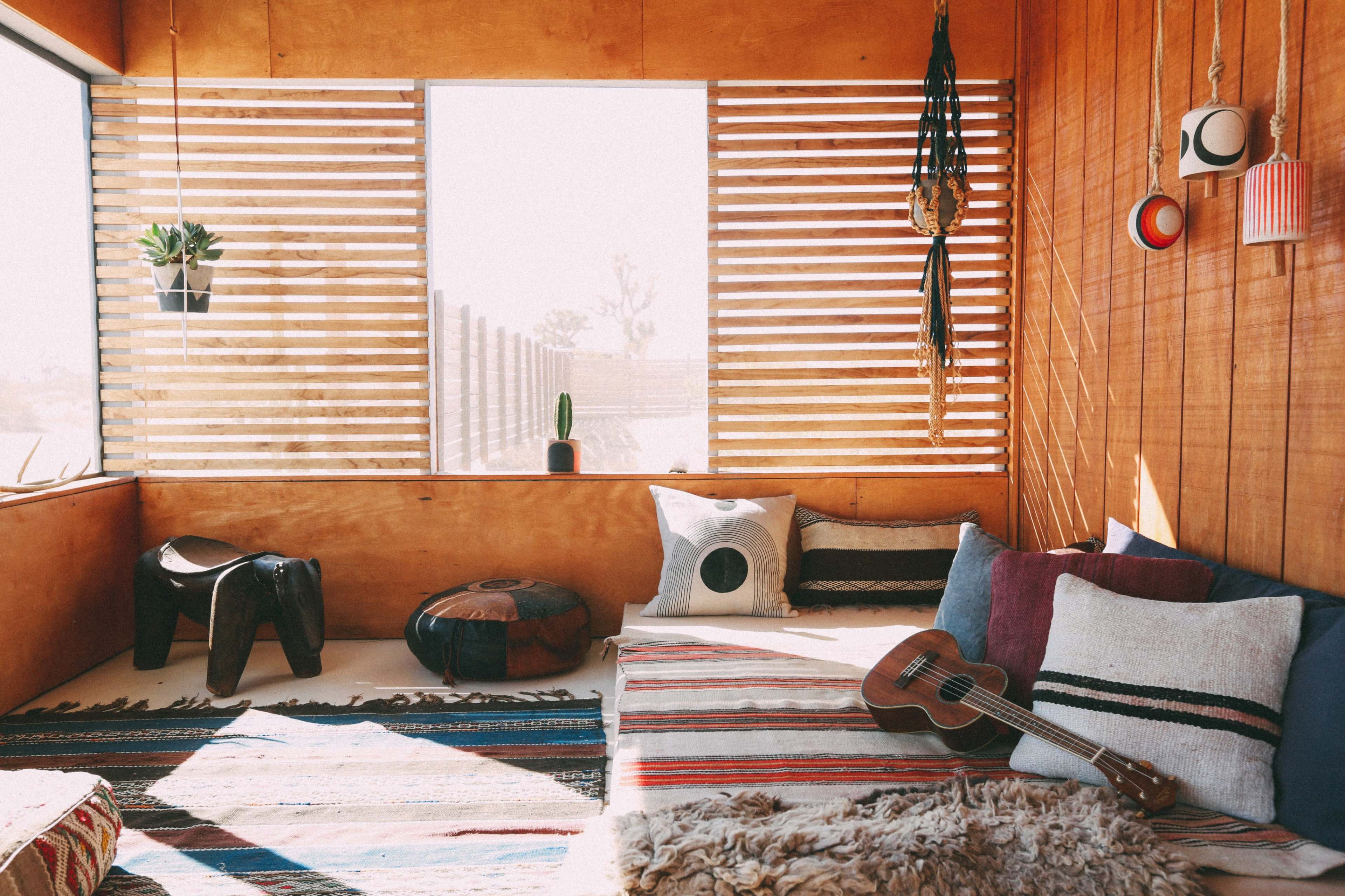 The scene features a cozy interior with wooden walls, a seating area adorned with patterned rugs and pillows, a ukulele resting nearby, and large windows allowing natural light to enter.