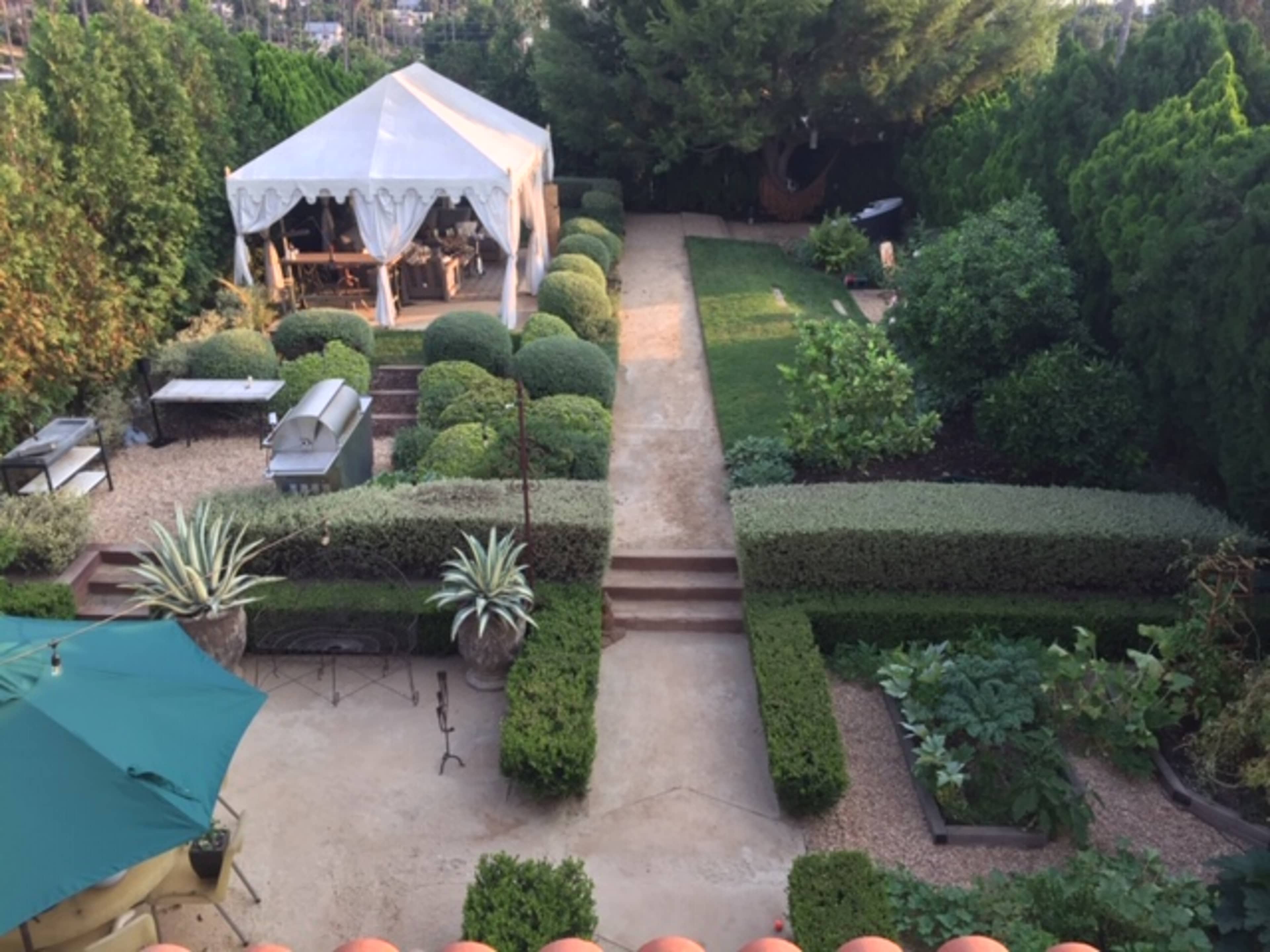 The image shows a landscaped garden featuring a pathway, neatly trimmed hedges, a tent, and various plants.