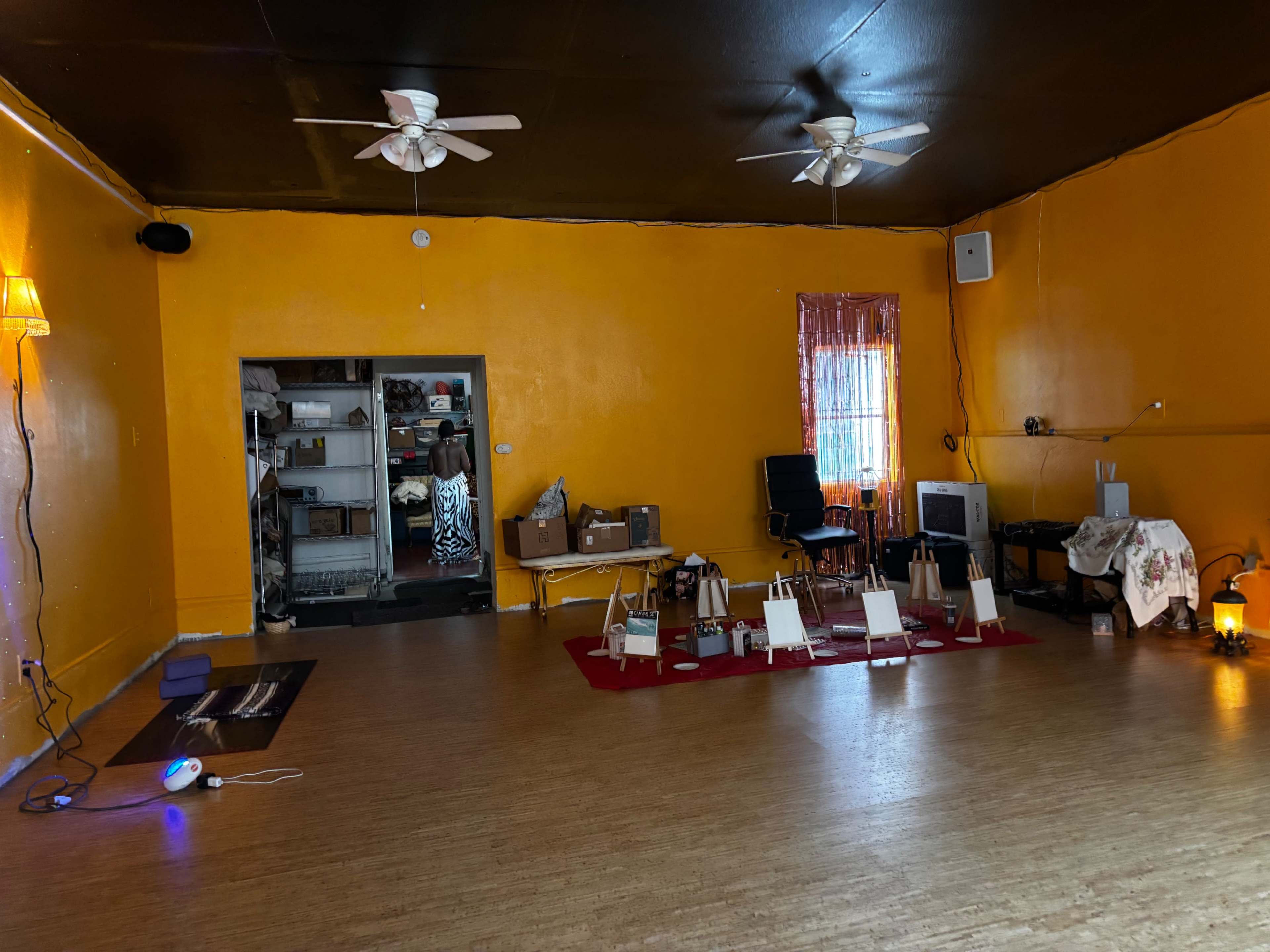 The image shows a spacious room with orange walls, hardwood flooring, two ceiling fans, and several easels arranged on a red carpet.
