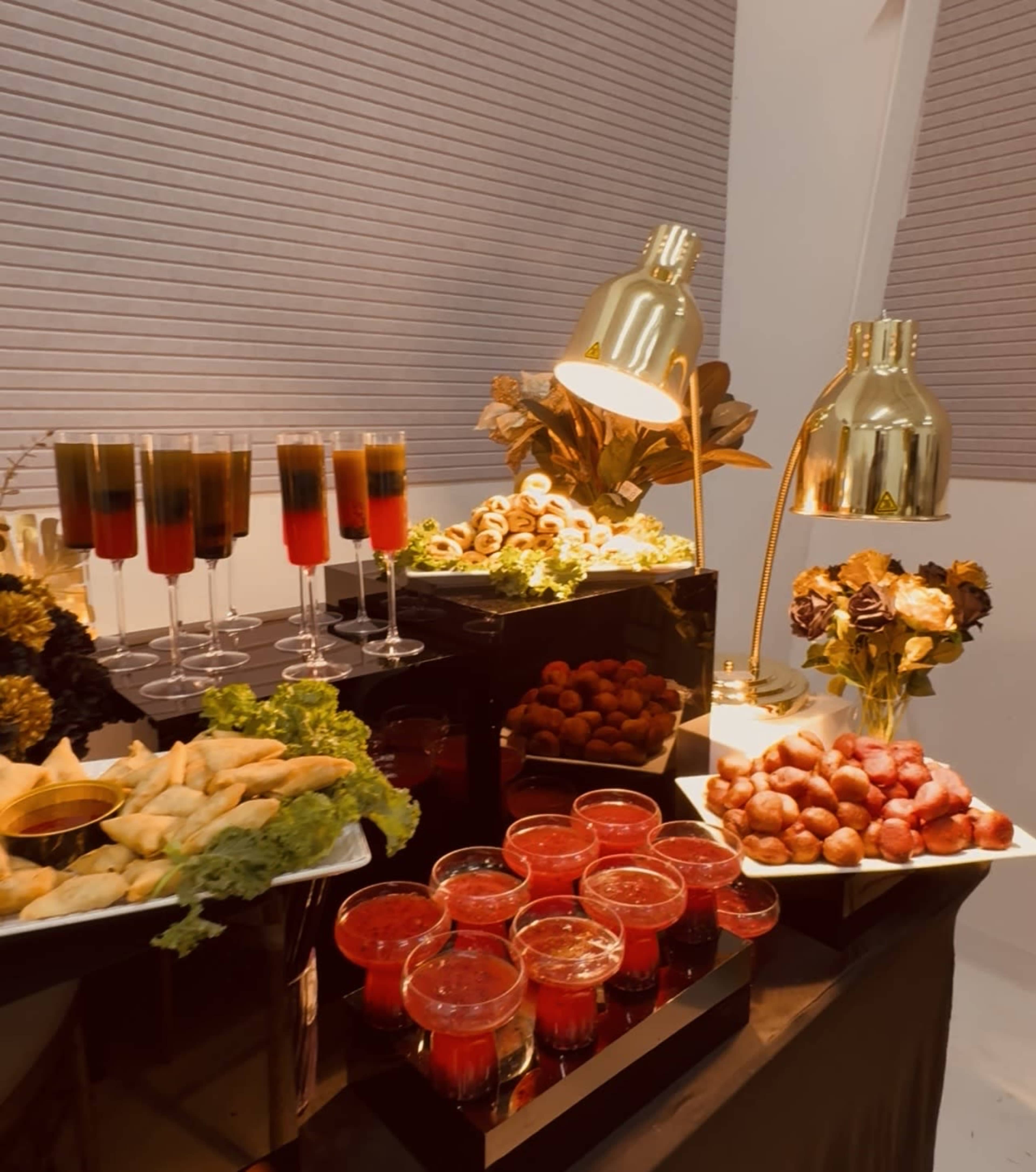 A stylish buffet display features an assortment of drinks in tall glasses, various appetizers on trays, and decorative lighting elements.
