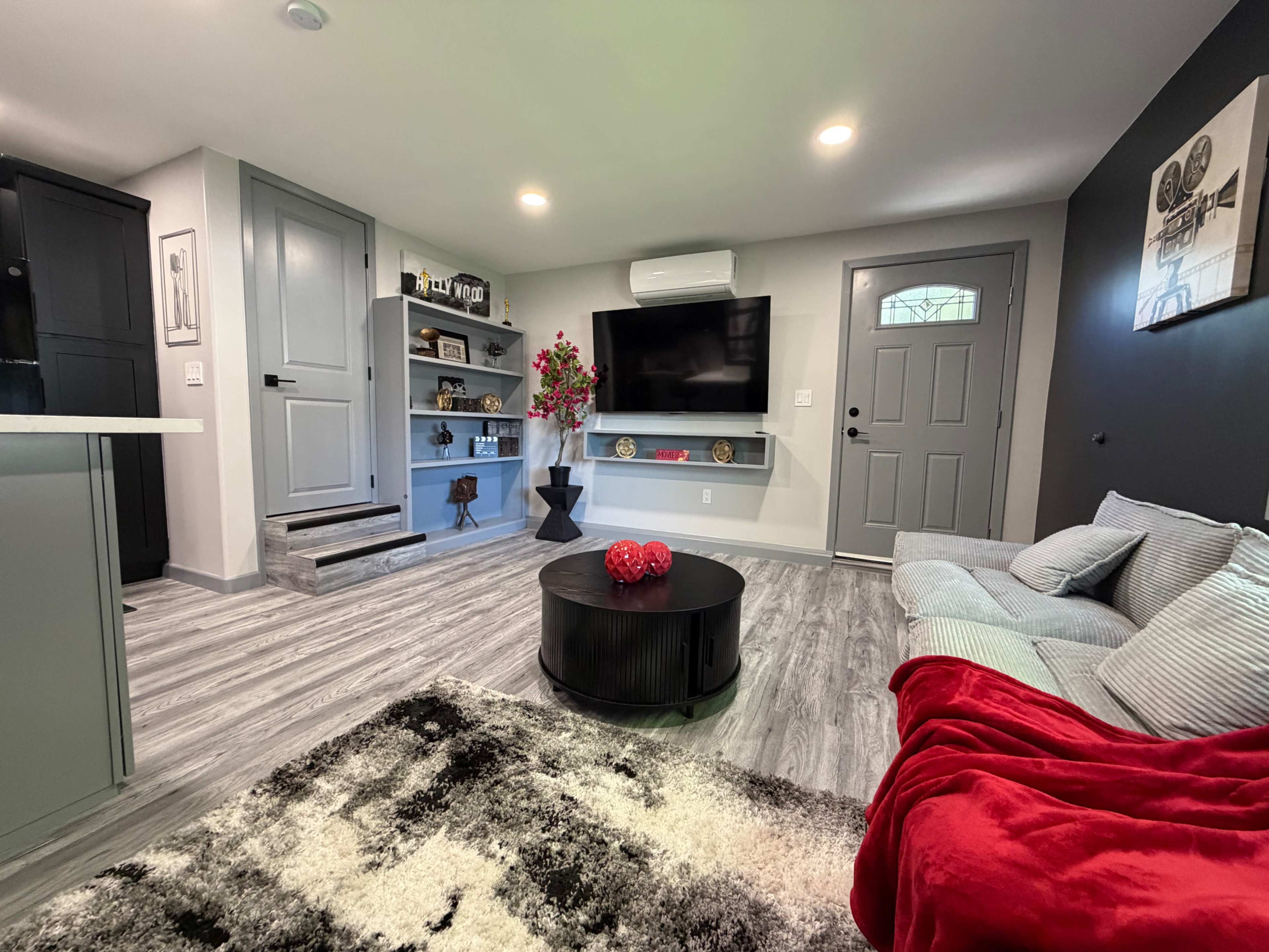 The image shows a modern living room featuring a gray sectional sofa, a black round coffee table, decorative shelves with plants and knick-knacks, and a TV mounted on the wall.