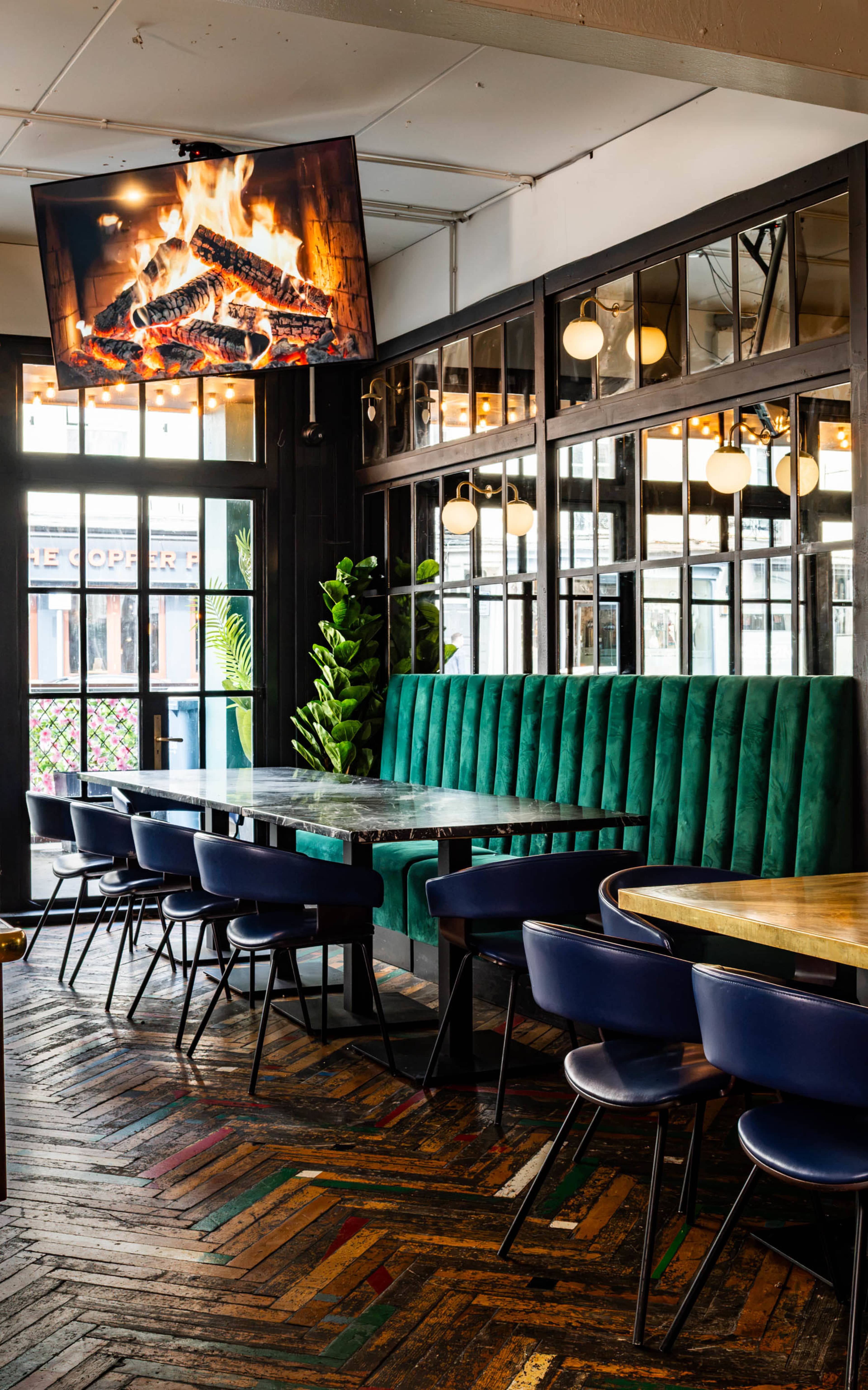The image shows a modern restaurant interior featuring green plush seating, wooden tables, and a wall-mounted television displaying a fireplace.
