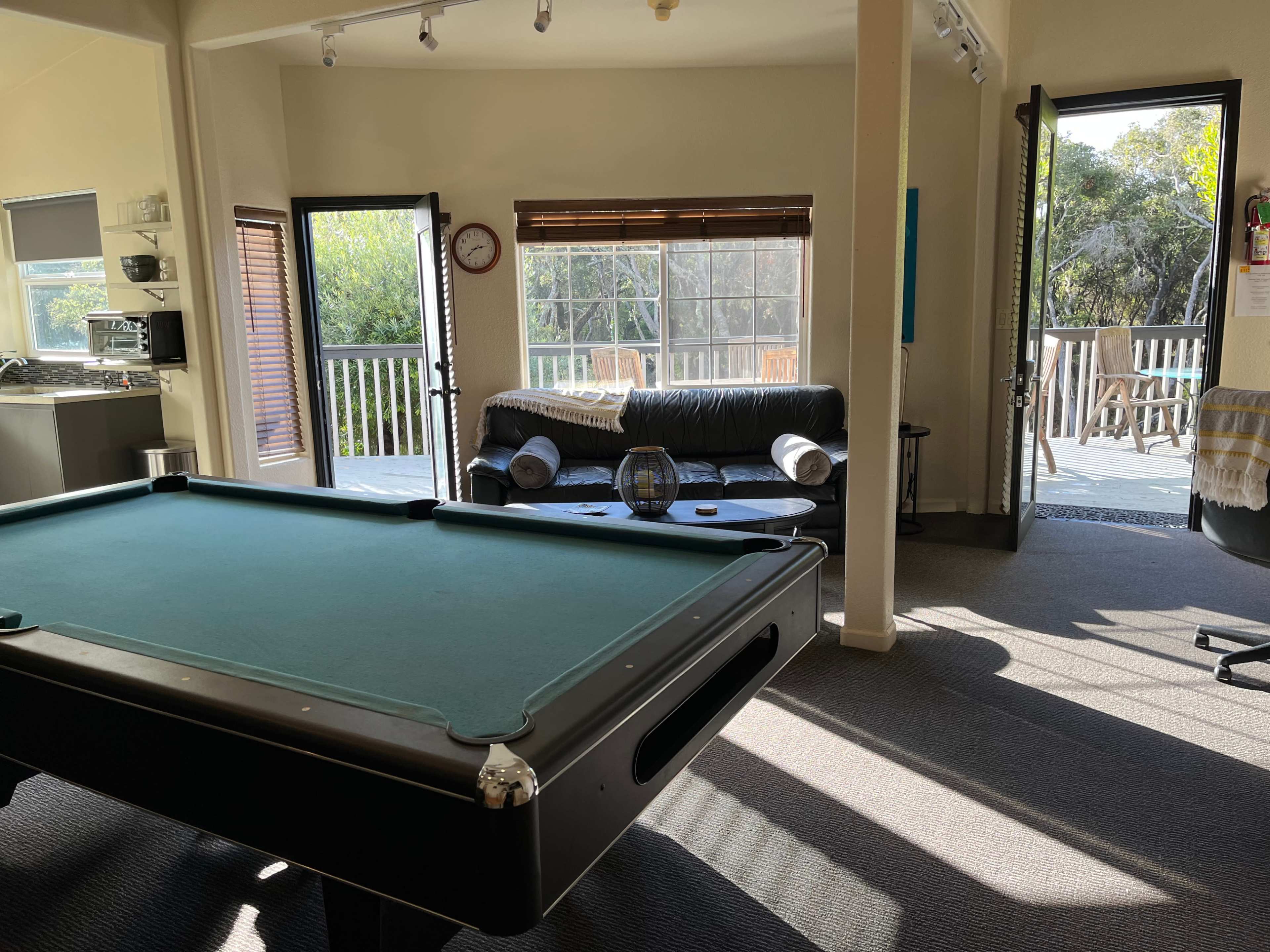 A sunlit room featuring a pool table, a leather couch, and large windows opening to an outdoor deck.