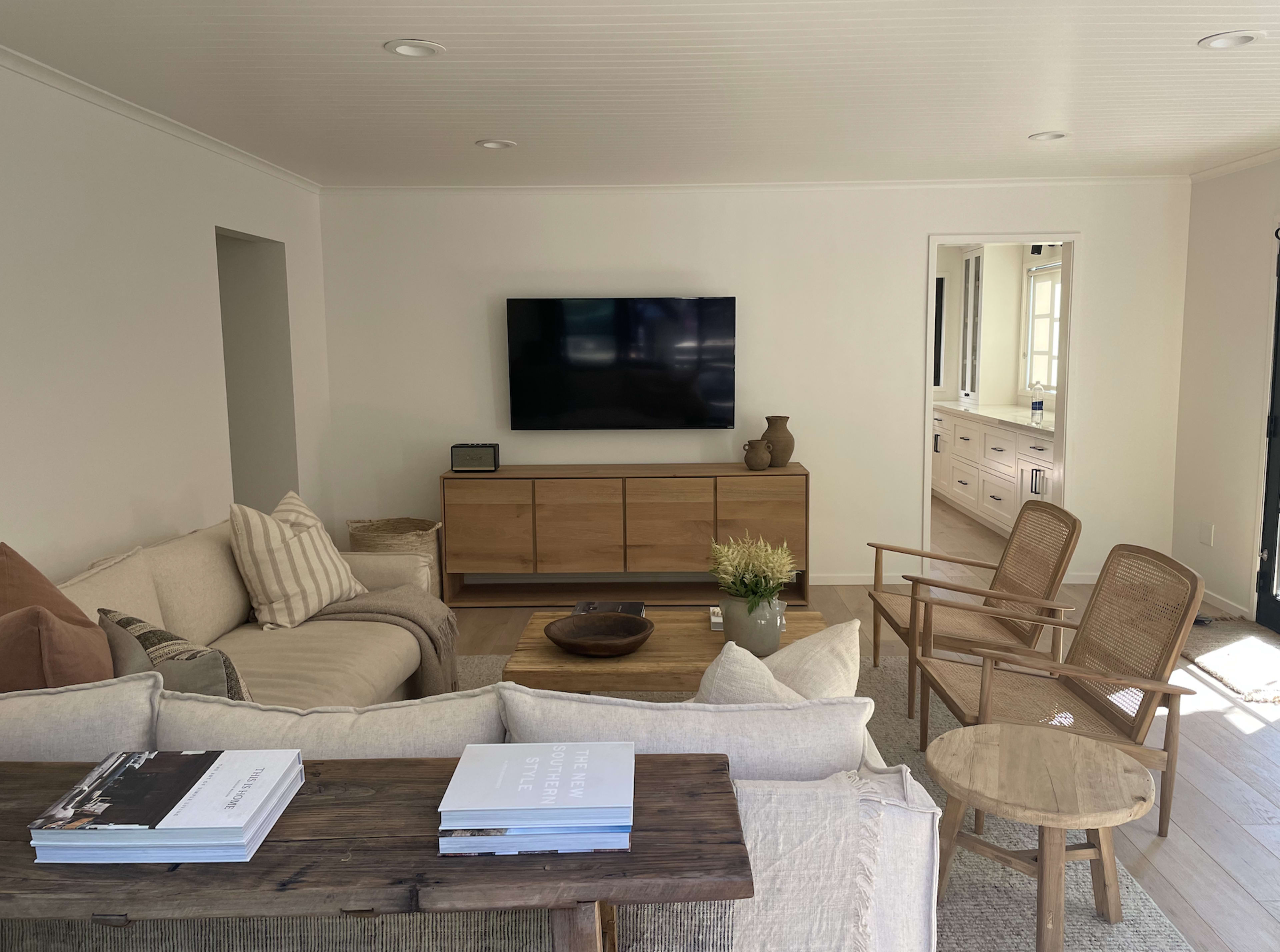 The living room features a couch with various cushions, a wooden coffee table, two chairs, a television mounted on the wall, and a view into a bathroom beyond.