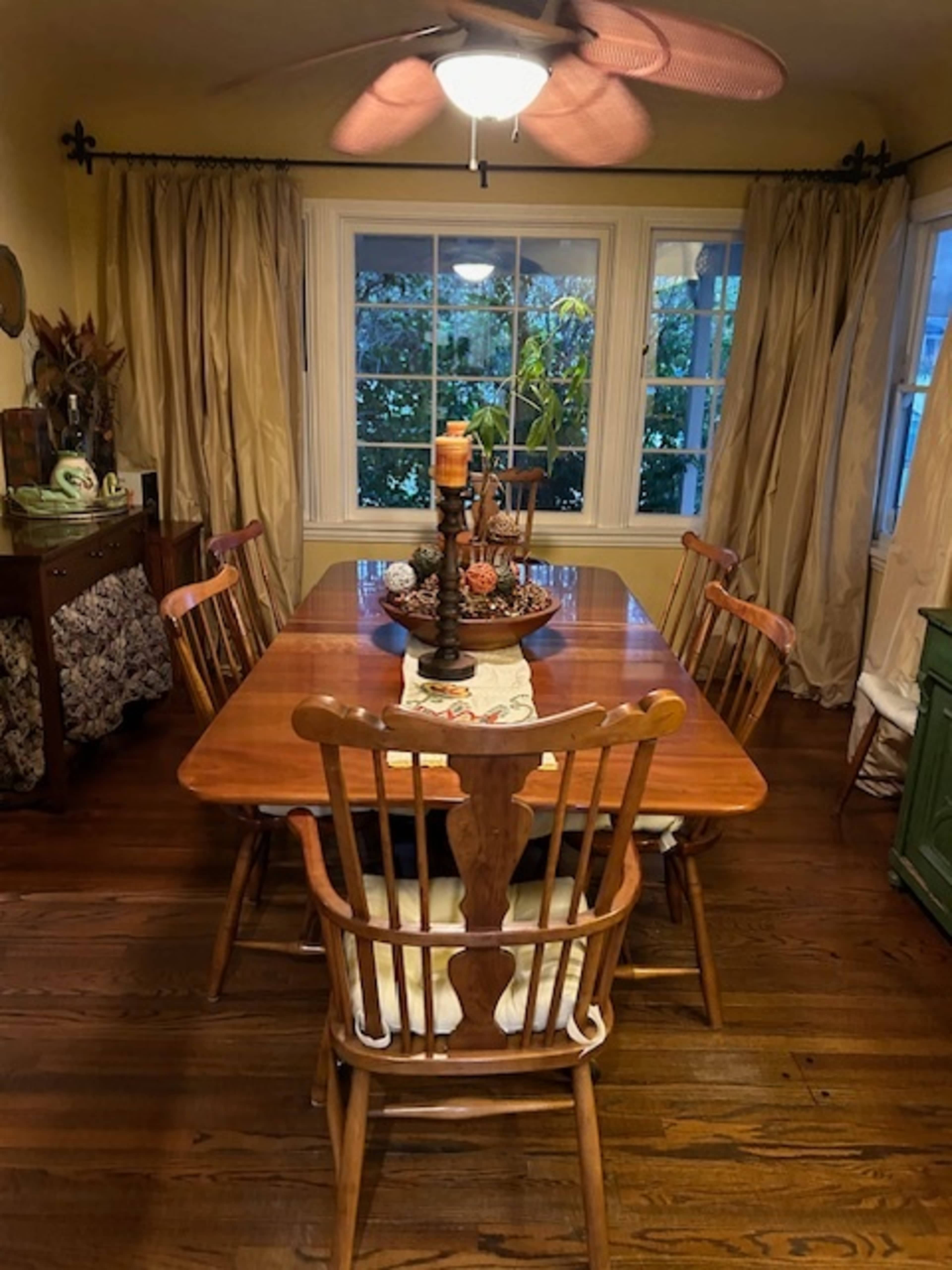 A wooden dining table with four chairs and a candle centerpiece is set in a room with large windows and draped curtains.
