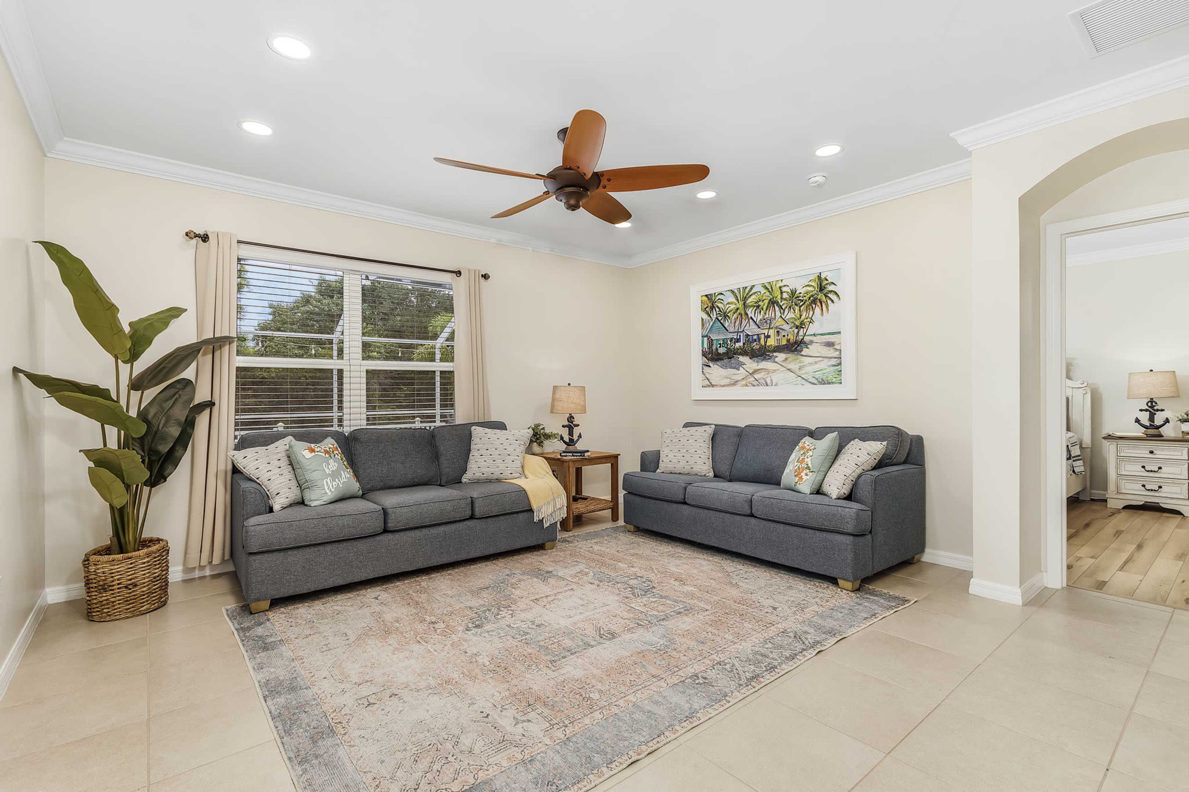 A cozy living room features two gray sofas, a patterned rug, and a palm plant, with soft lighting and artwork on the wall.