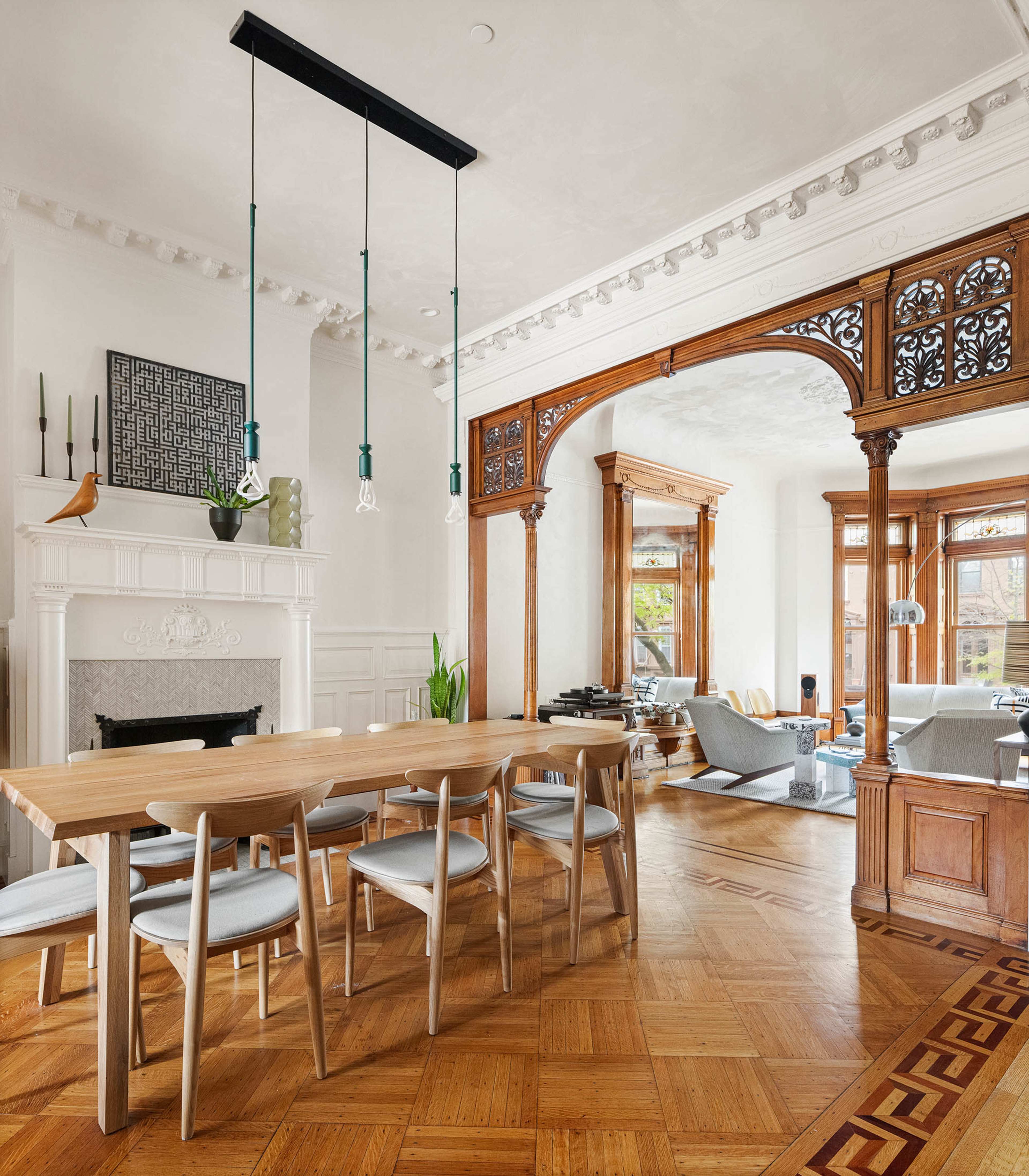 A wooden dining table with six chairs is positioned under pendant lights in a room featuring ornate woodwork and a fireplace, with a living area visible in the background.